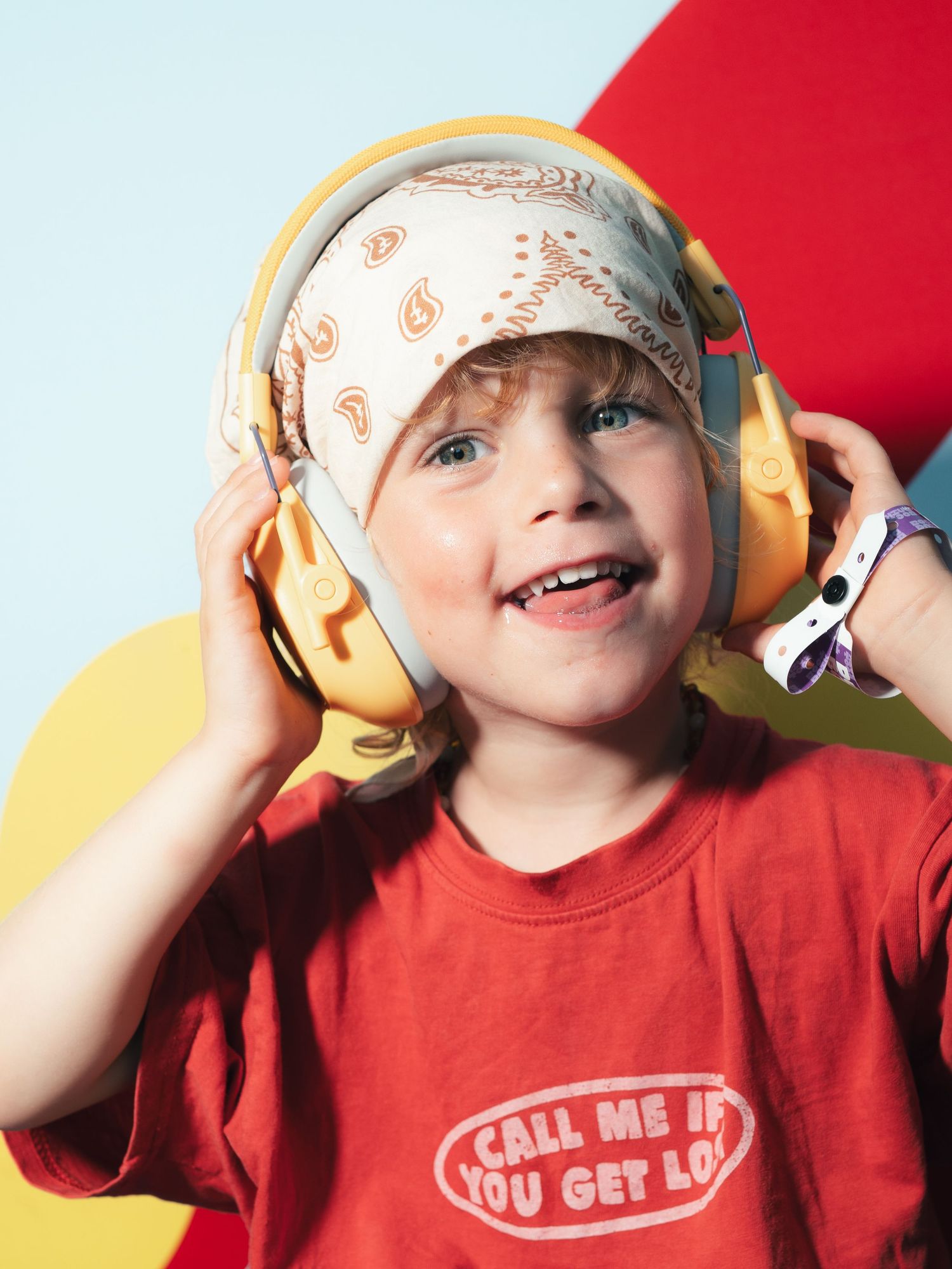 Happy child wearing yellow headphones and a red t-shirt with a bandana on their head against a colorful backdrop.