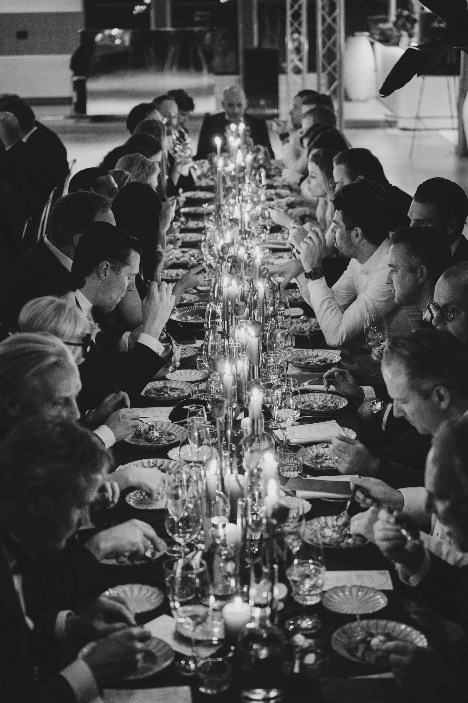 Black and white photo of elegantly dressed guests dining at a long candlelit banquet table.