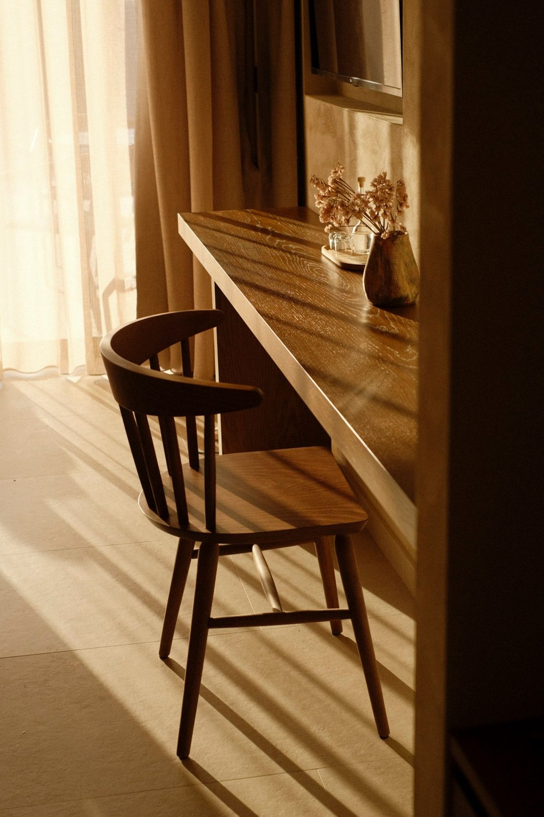 Warm sunlight casts shadows on a wooden chair beside a narrow desk with dried flowers in a cozy room.