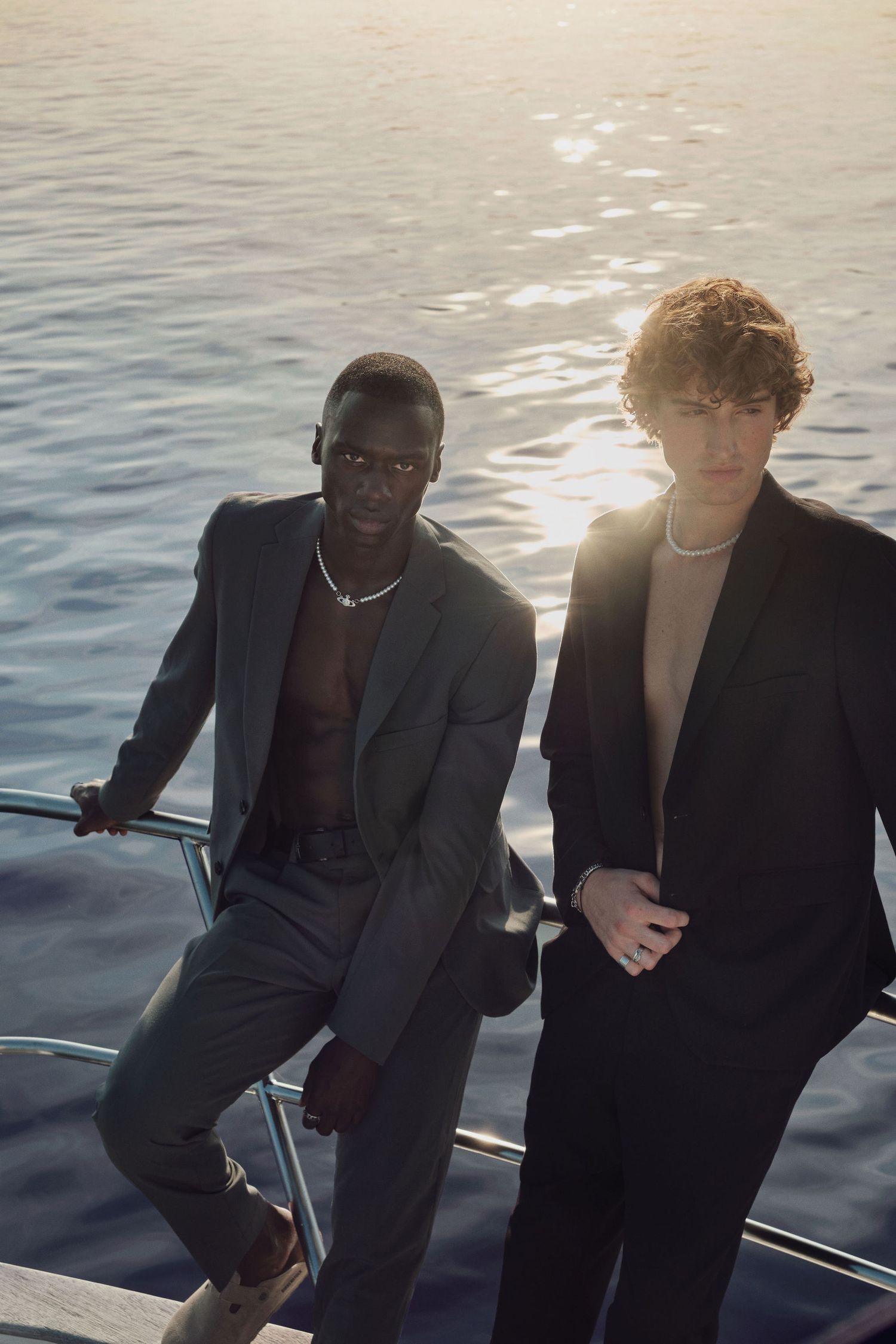 Two stylish men in dark suits pose on a yacht at sunset over shimmering ocean waters.