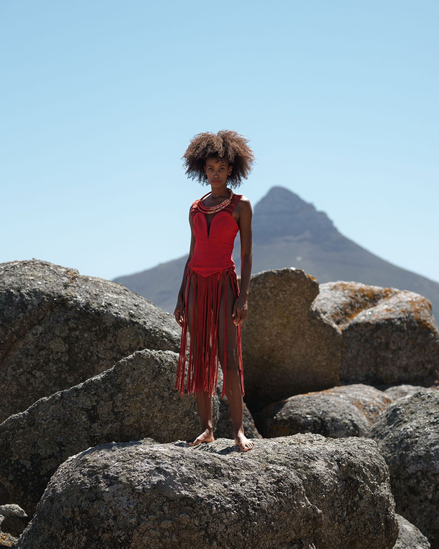 Woman in red fringe dress standing on rocks with mountain backdrop under clear blue sky.