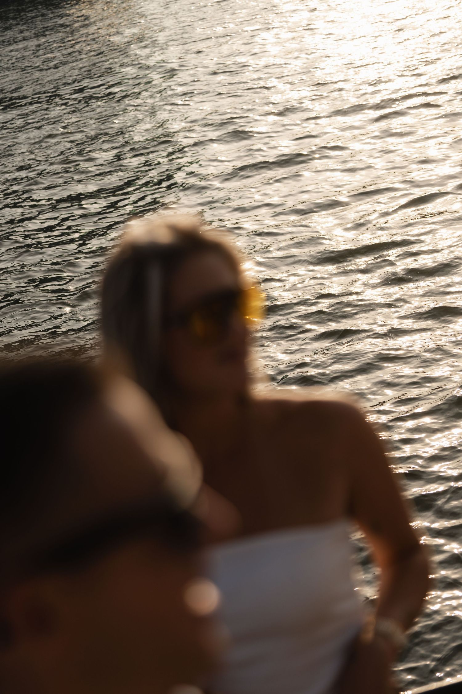 A blond woman wearing sunglasses relaxes on a boat at sunset with golden light reflecting on the water.