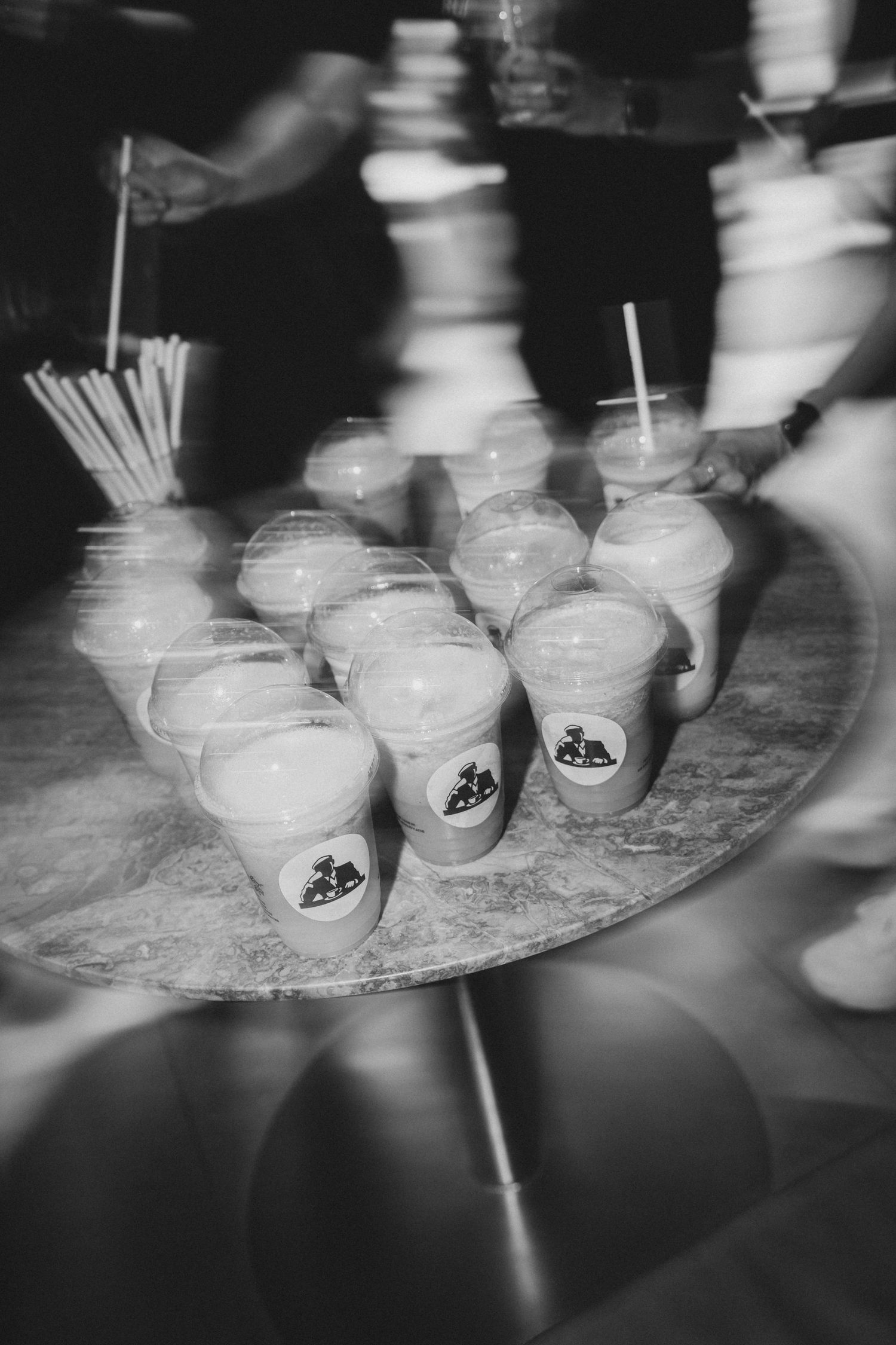 Black and white photo of branded plastic cups with straws arranged on a round tray at an event.