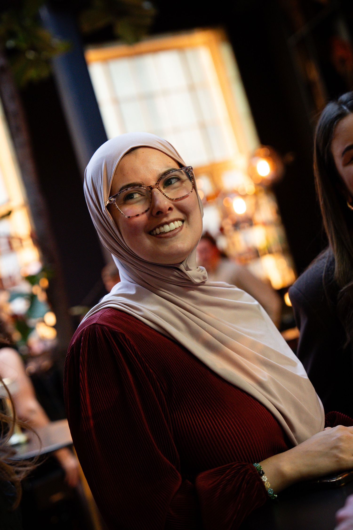 Person wearing glasses and white hijab smiling warmly in an indoor setting with warm lighting.