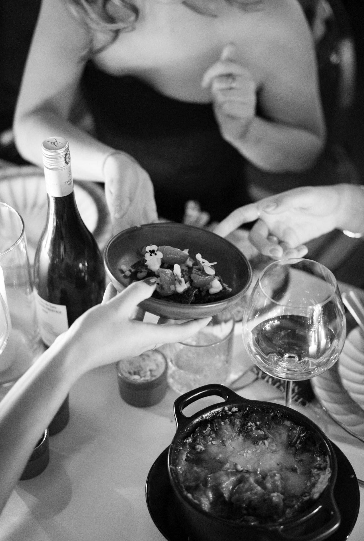 Black and white close-up of hands passing a bowl of food at an elegant dinner with wine and candles.