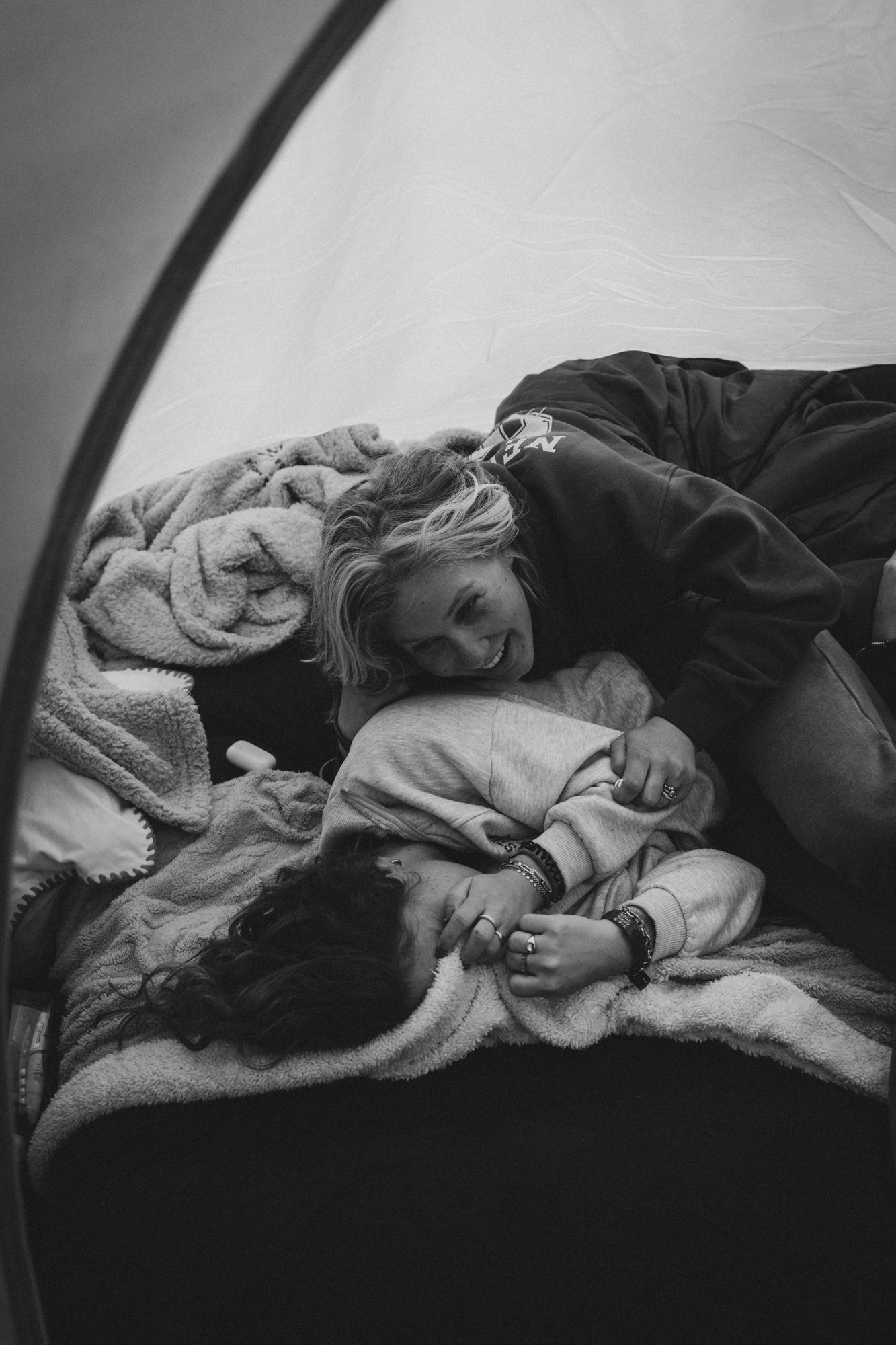 Black and white photo of two women cuddling together under blankets inside a cozy camping tent.