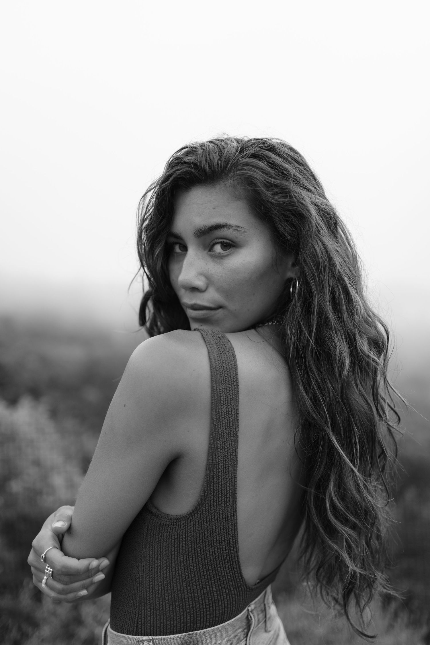 Black and white portrait of a young woman with long wavy hair in a backless top, looking over her shoulder outdoors.
