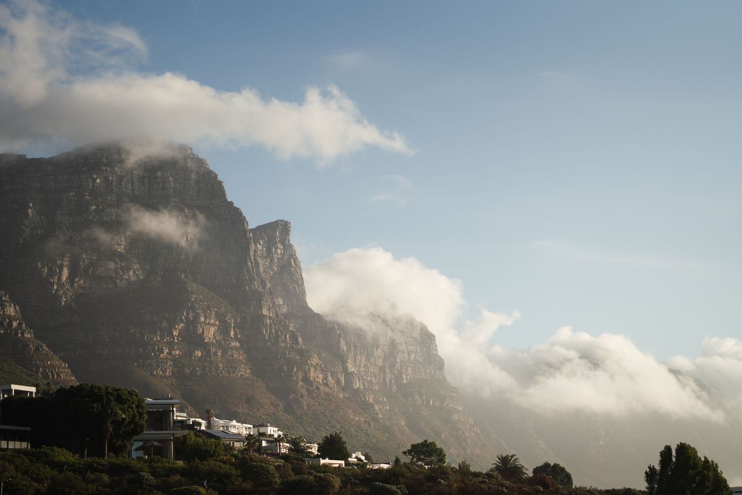 Dramatic rocky mountain cliffs with swirling clouds above a small village, Cape Town's Table Mountain landscape.