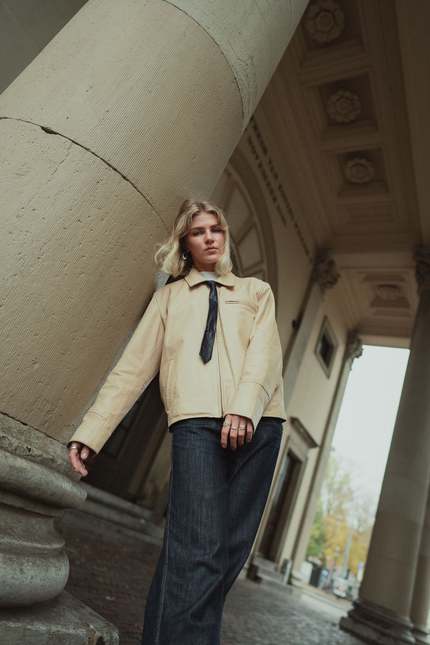 Blonde woman in beige leather jacket and dark trousers with tie poses between classical stone columns.