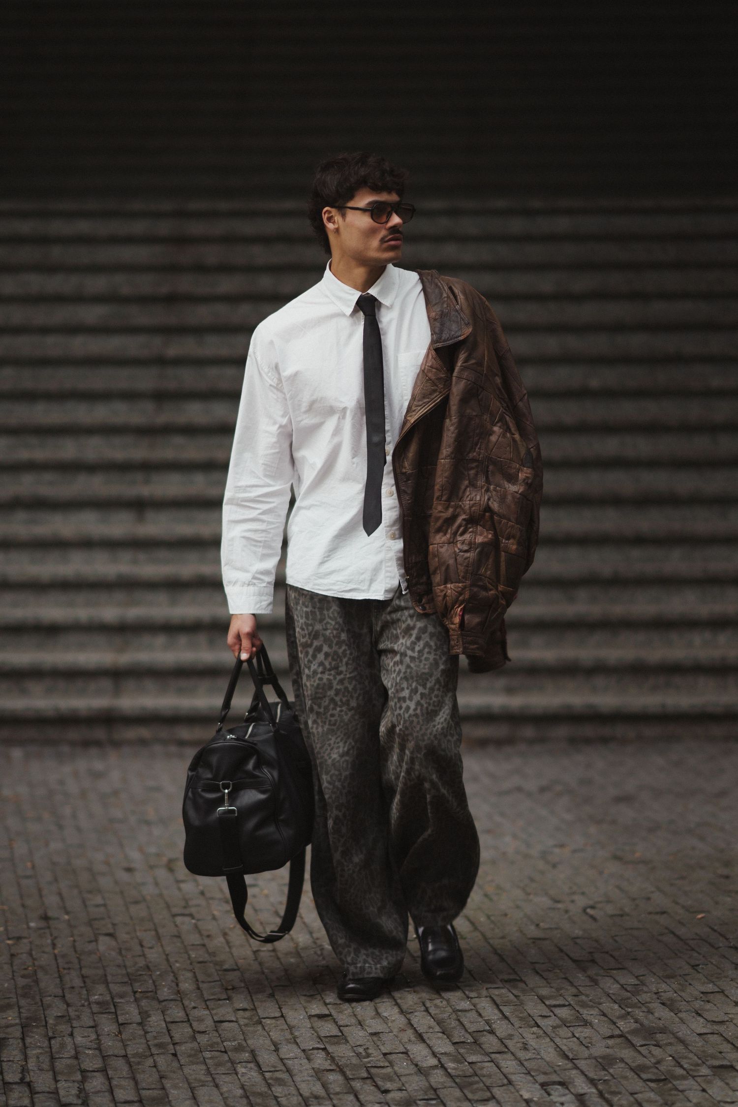 Man in white shirt, black tie, brown leather jacket, gray pants, and black bag walking on cobblestone.