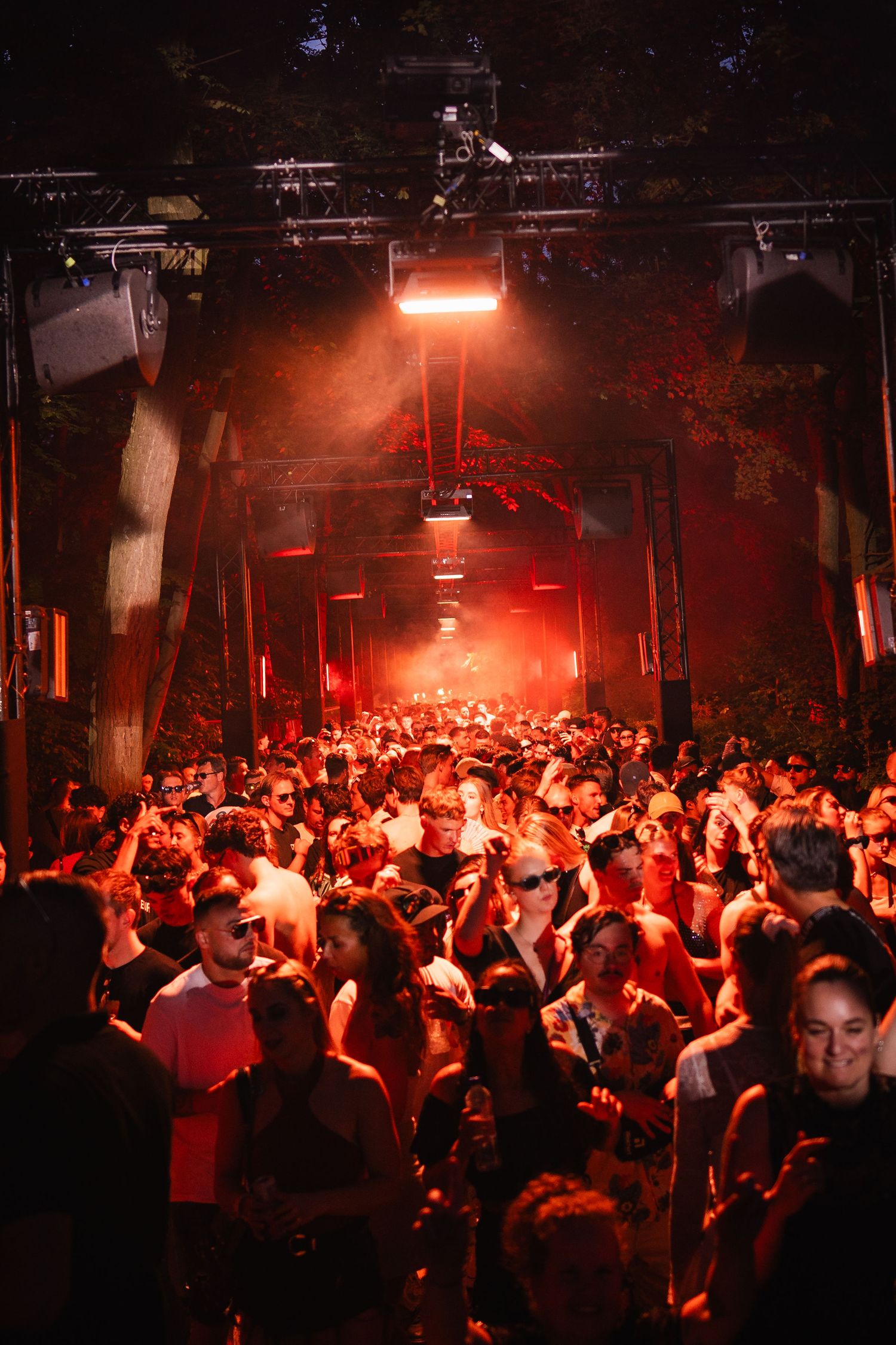A large crowd of people dancing under red lights at an outdoor nightclub or music festival event.