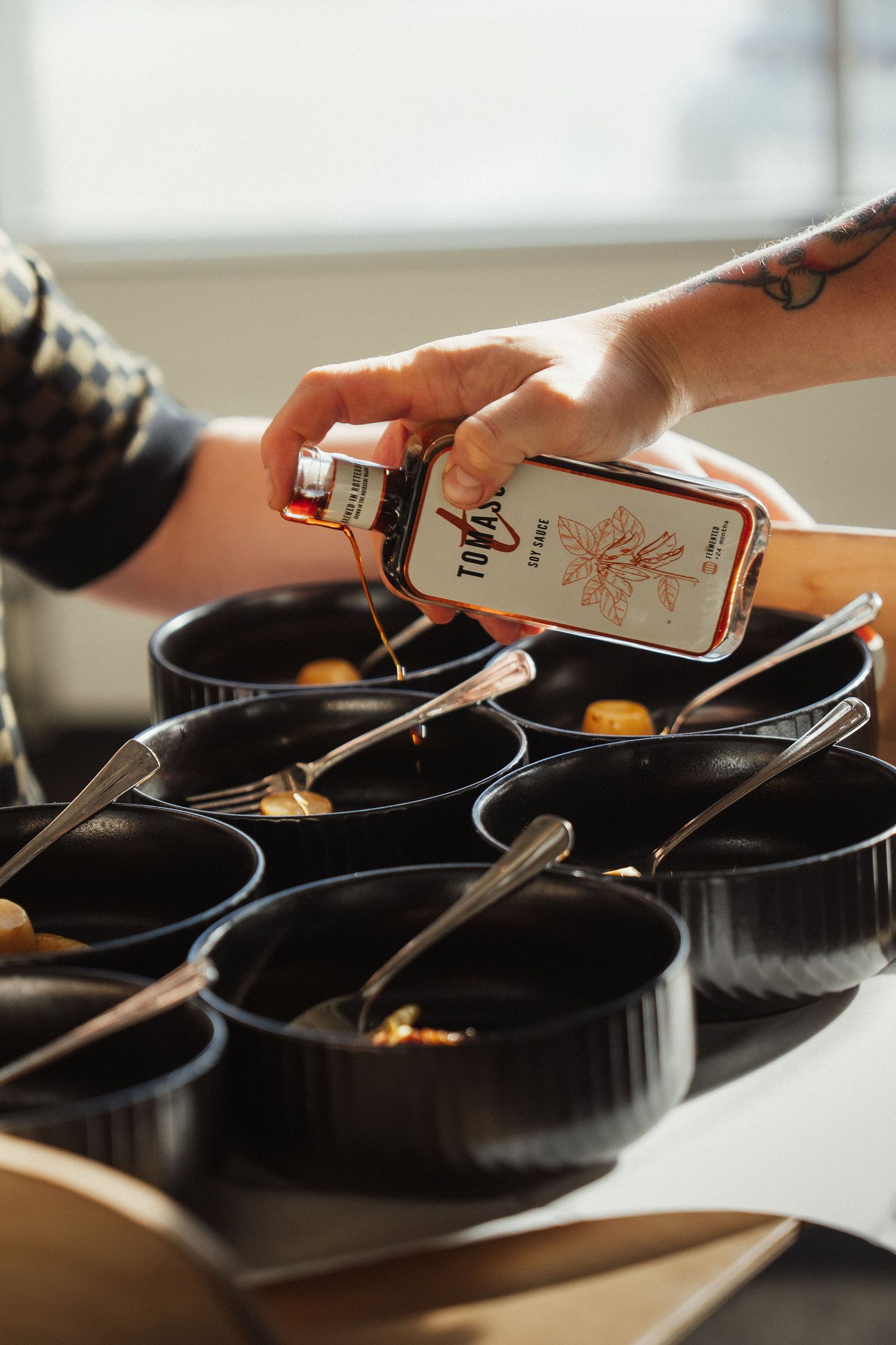 Hands pouring Tofol sauce into black bowls with spoons on a table in natural light.