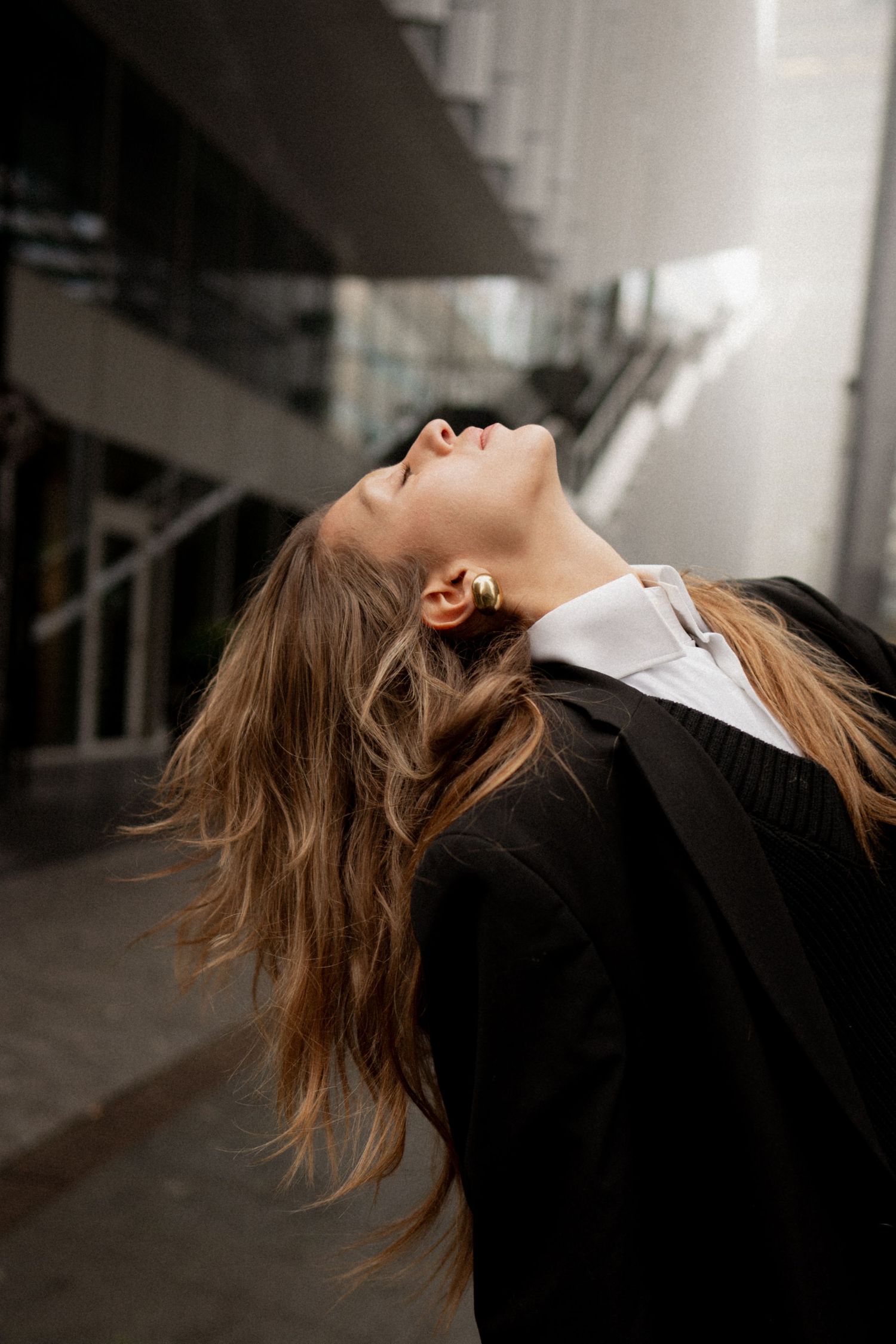 A woman in a black blazer tilts her head back with flowing hair outdoors near a modern building.