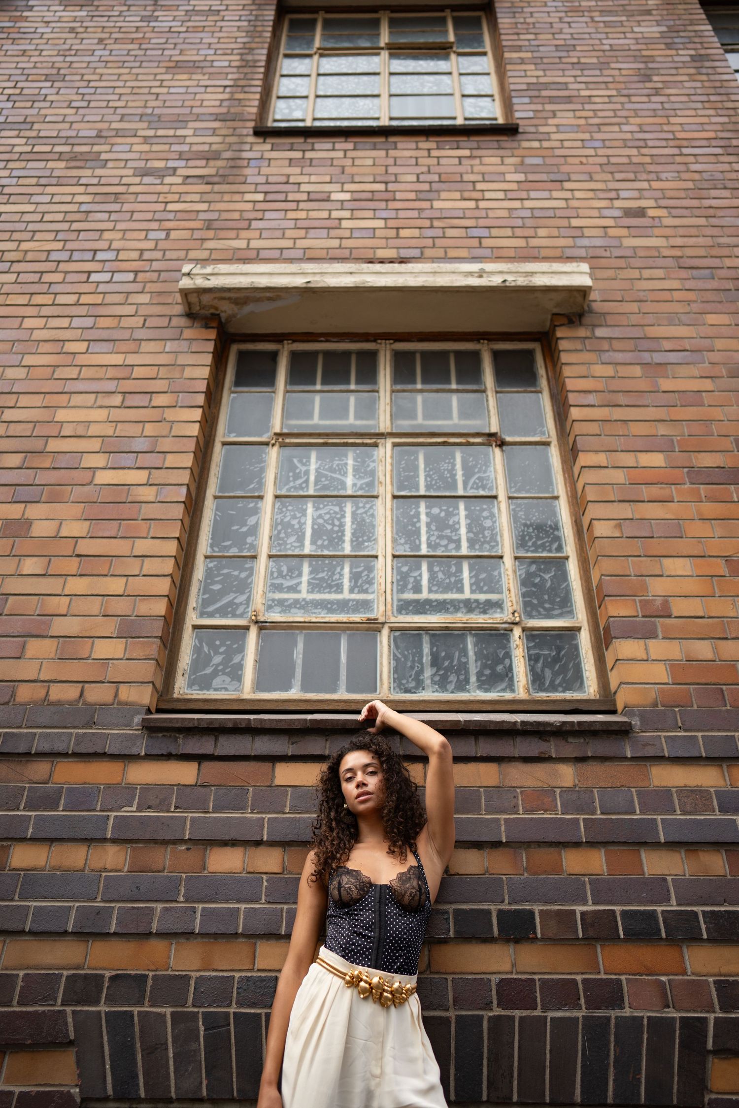 Young woman with curly hair posing against a vintage brick building wall with large industrial windows.