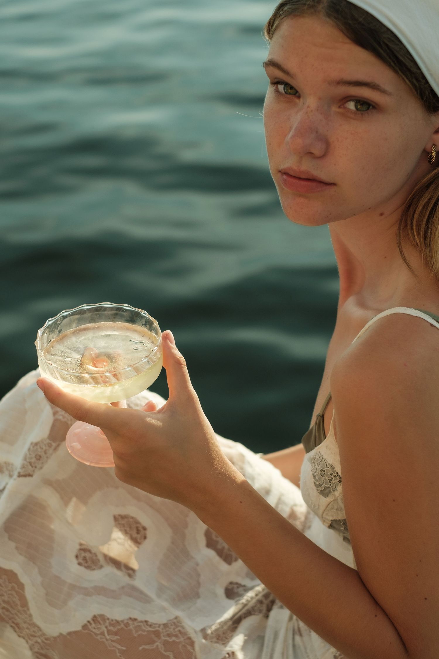Young woman in white lace dress holding a cocktail glass near the ocean, looking at camera.
