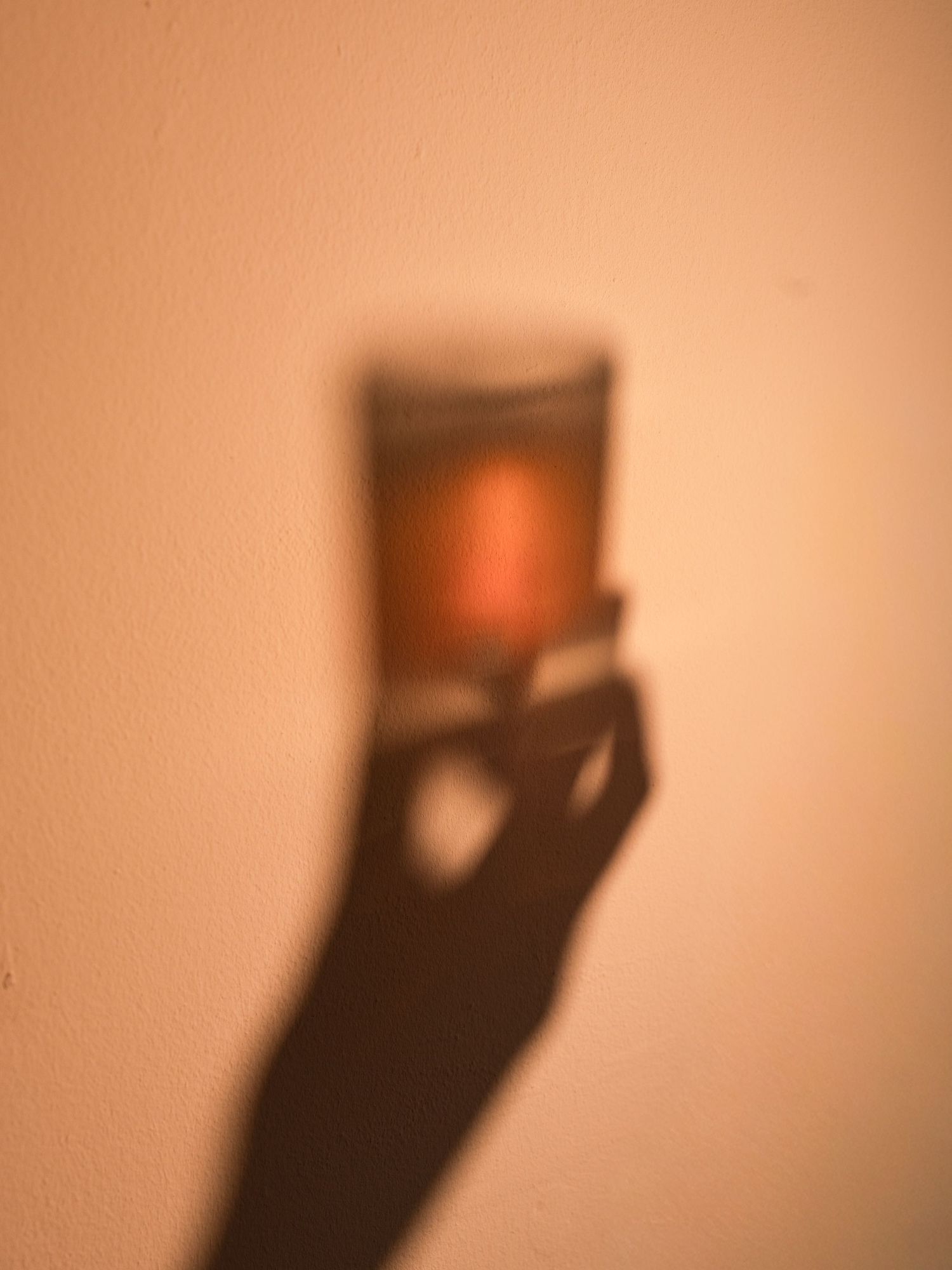 Shadow of a hand holding a glowing candle projected on a warm beige wall.