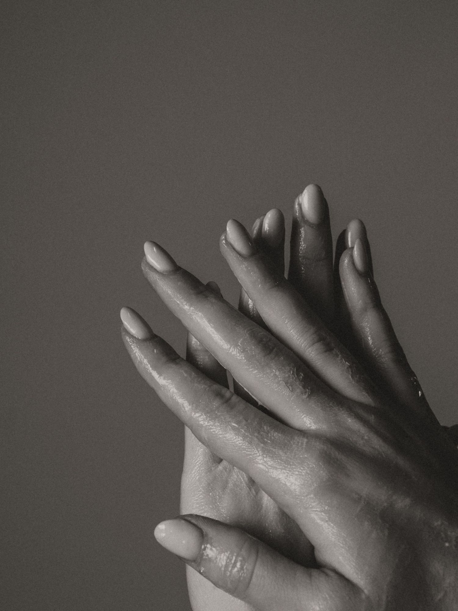 Black and white close-up of two hands with oval nails intertwined, glistening with moisture against a gray background.