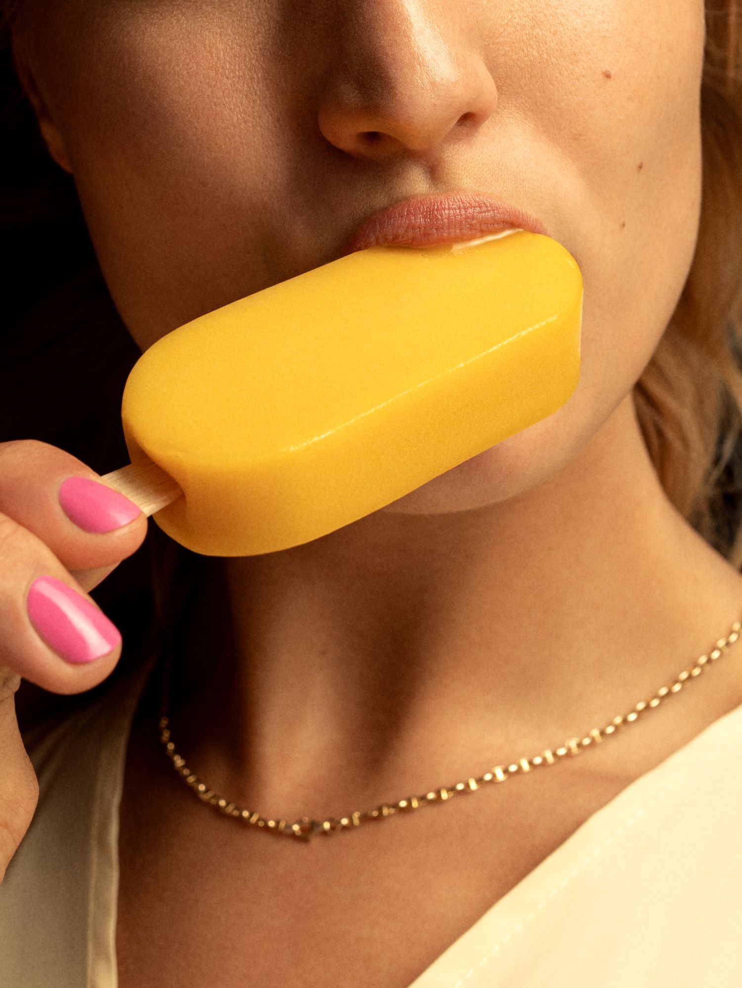 Woman with pink nails holding a yellow mango popsicle up to her lips, wearing a gold chain necklace.