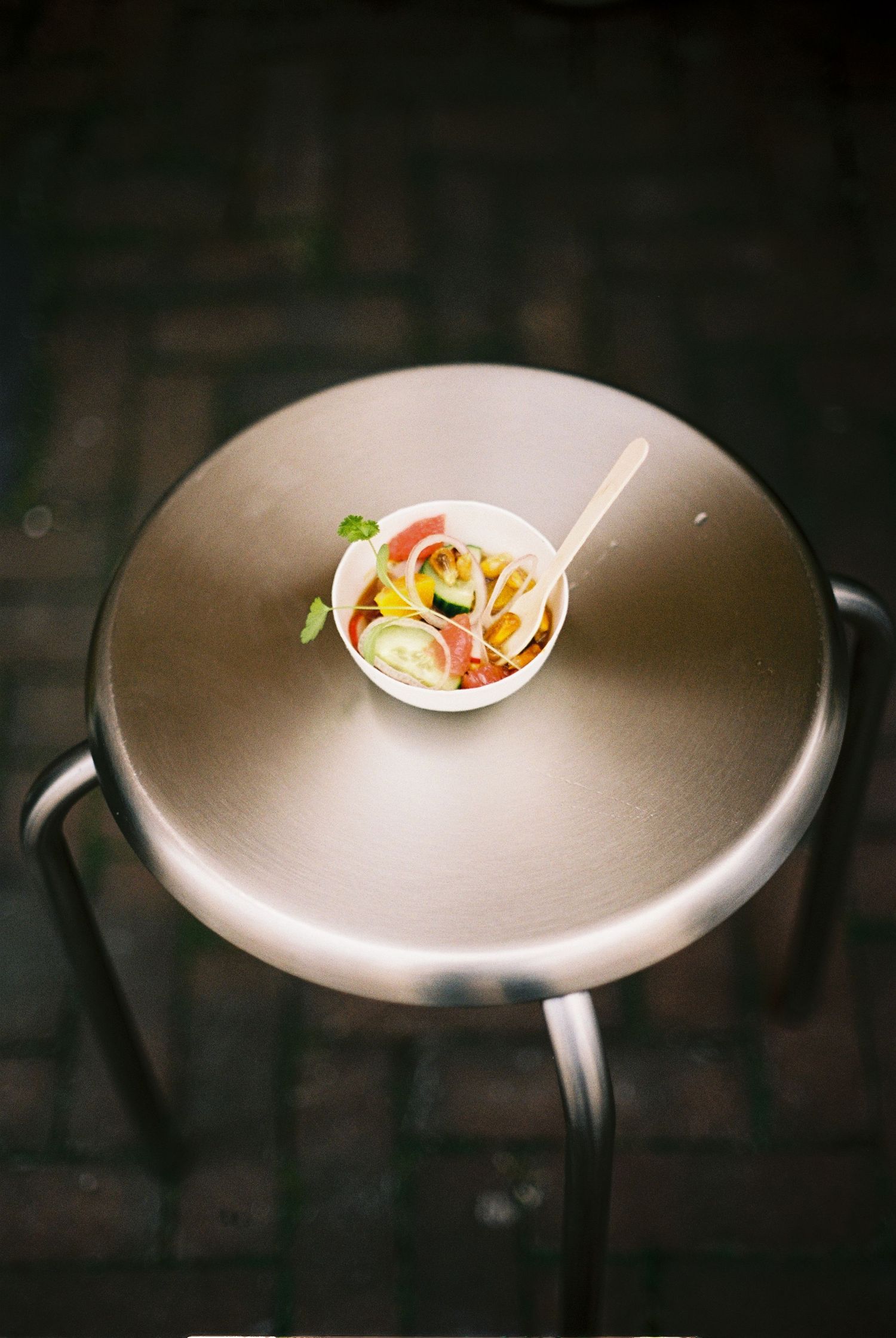 A small bowl of colorful salad with a wooden fork on a round silver stool outdoors on brick pavement.