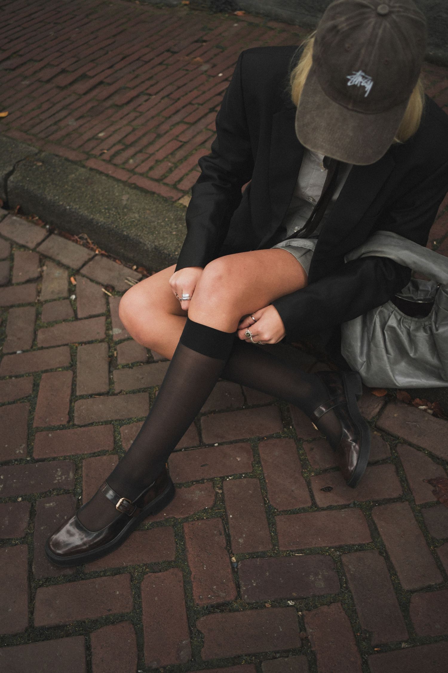 Woman in black blazer and knee-high socks adjusts stockings while seated on brick pavement.