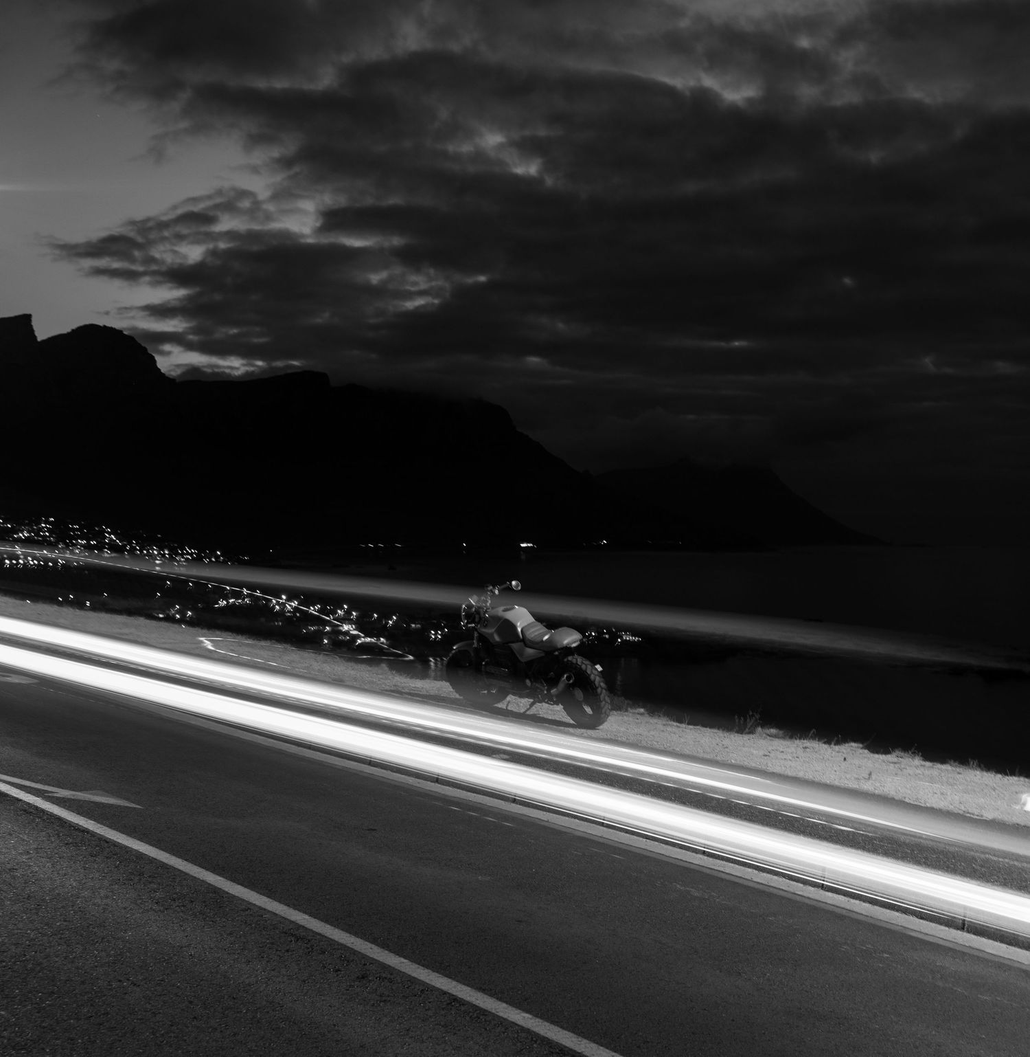 Black and white long exposure photo of a motorcycle on a coastal road at night with light trails and dramatic cloudy sky.