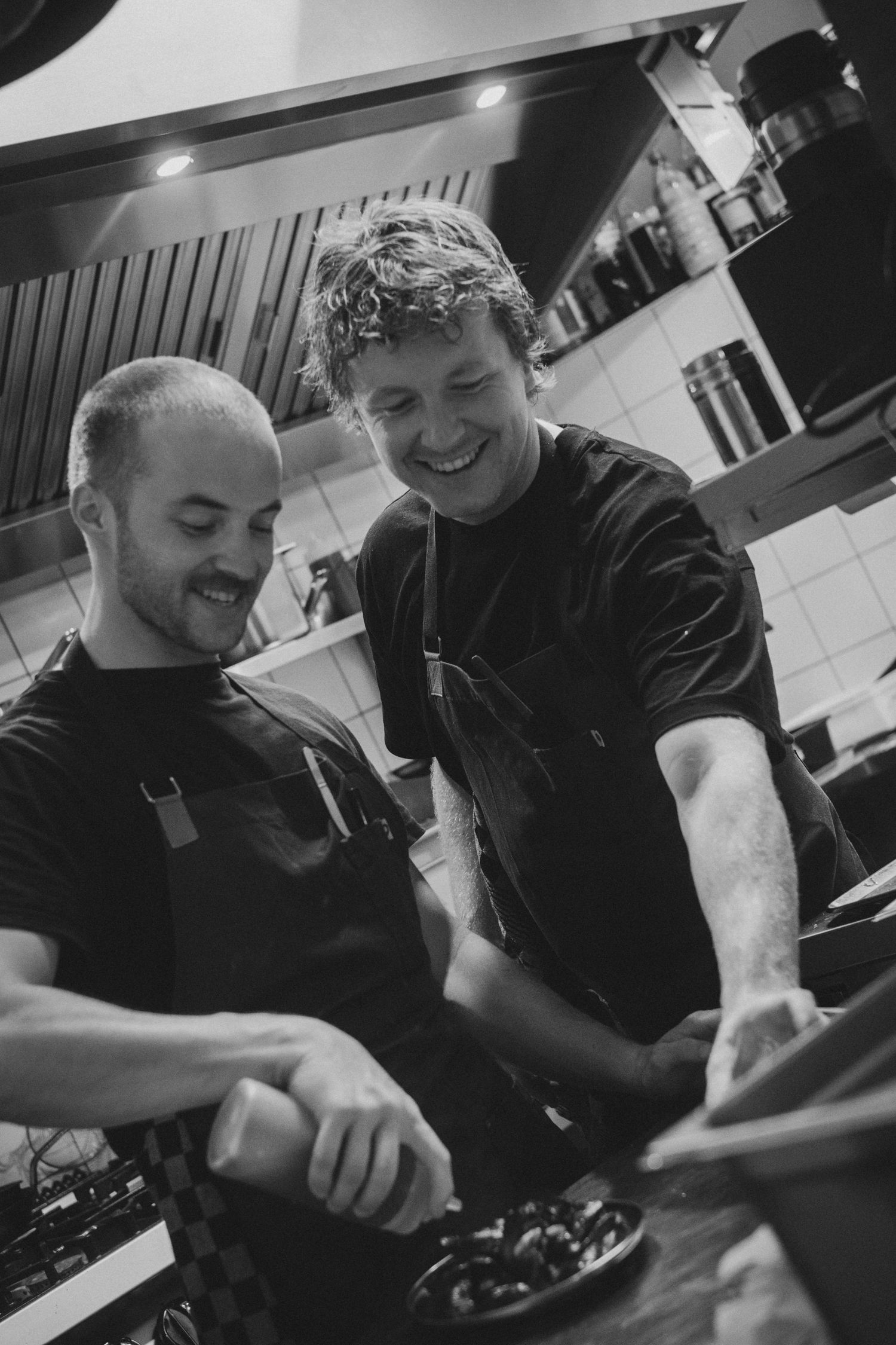 Two smiling chefs in black uniforms collaborate over a dish in a professional kitchen.