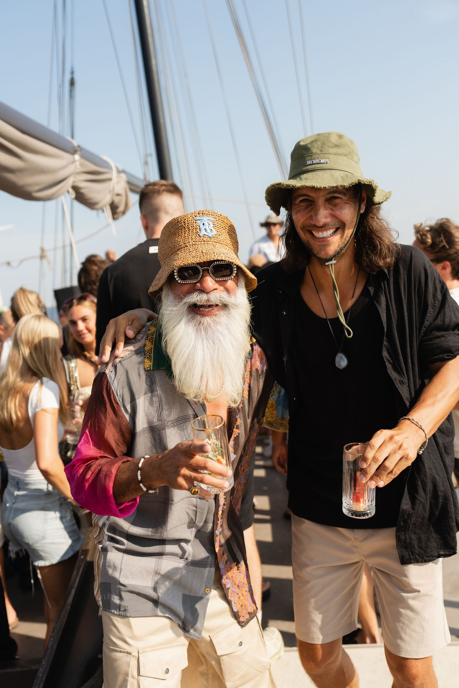 Two men wearing bucket hats smile and hold drinks on a sunny sailboat deck surrounded by guests.