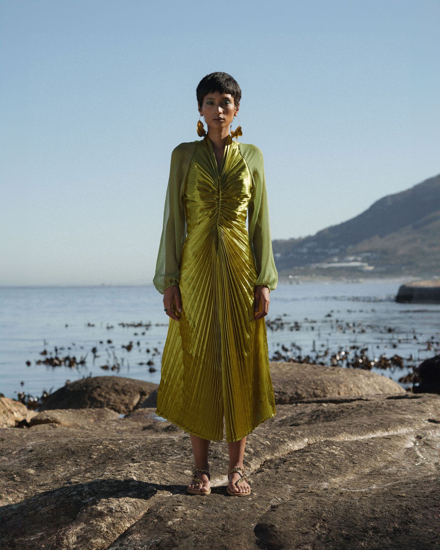 Woman in chartreuse pleated midi dress with sheer sleeves stands on rocky shore with ocean and mountain backdrop.