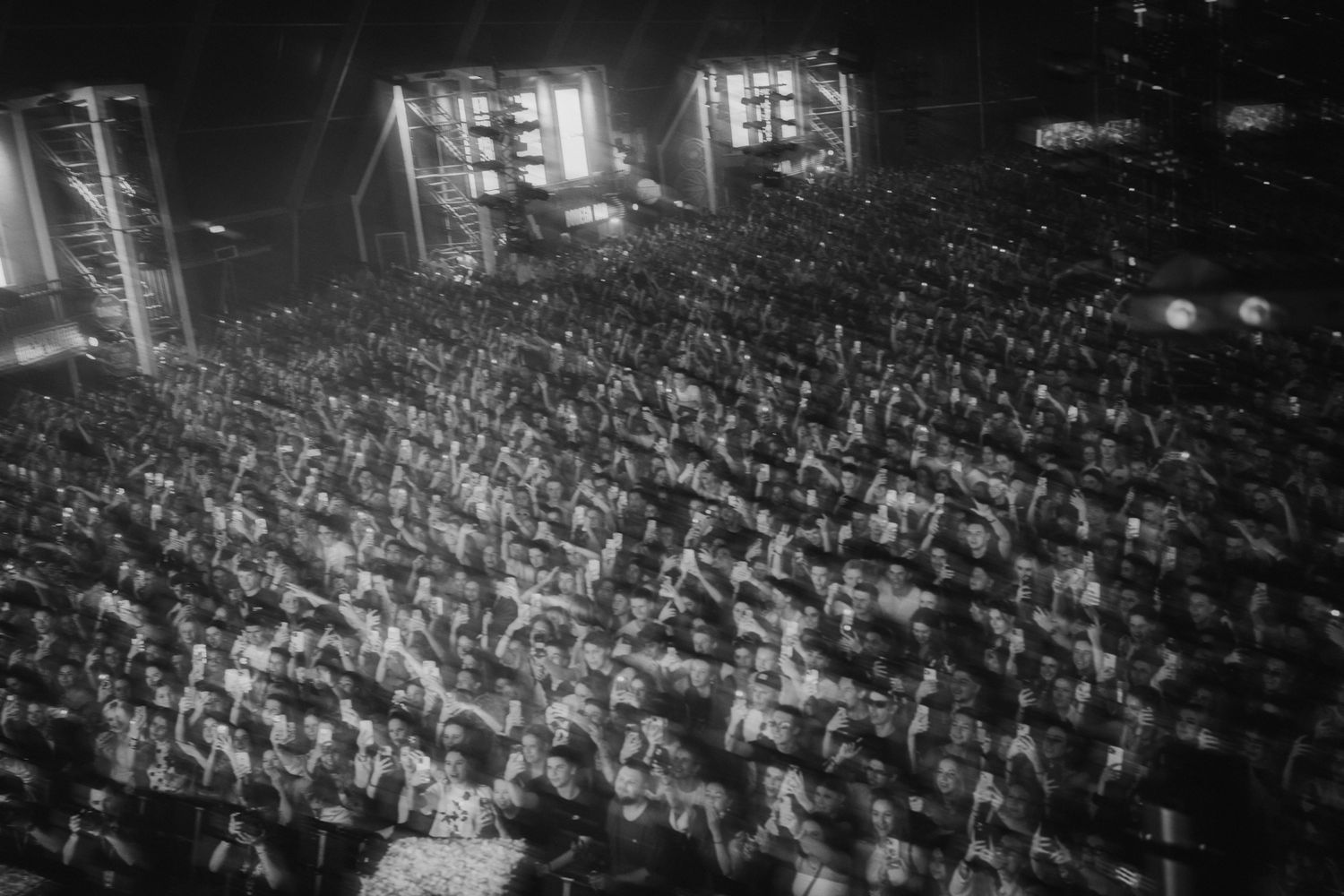 Black and white aerial view of a massive outdoor concert crowd at night with bright stage lights illuminating the scene.