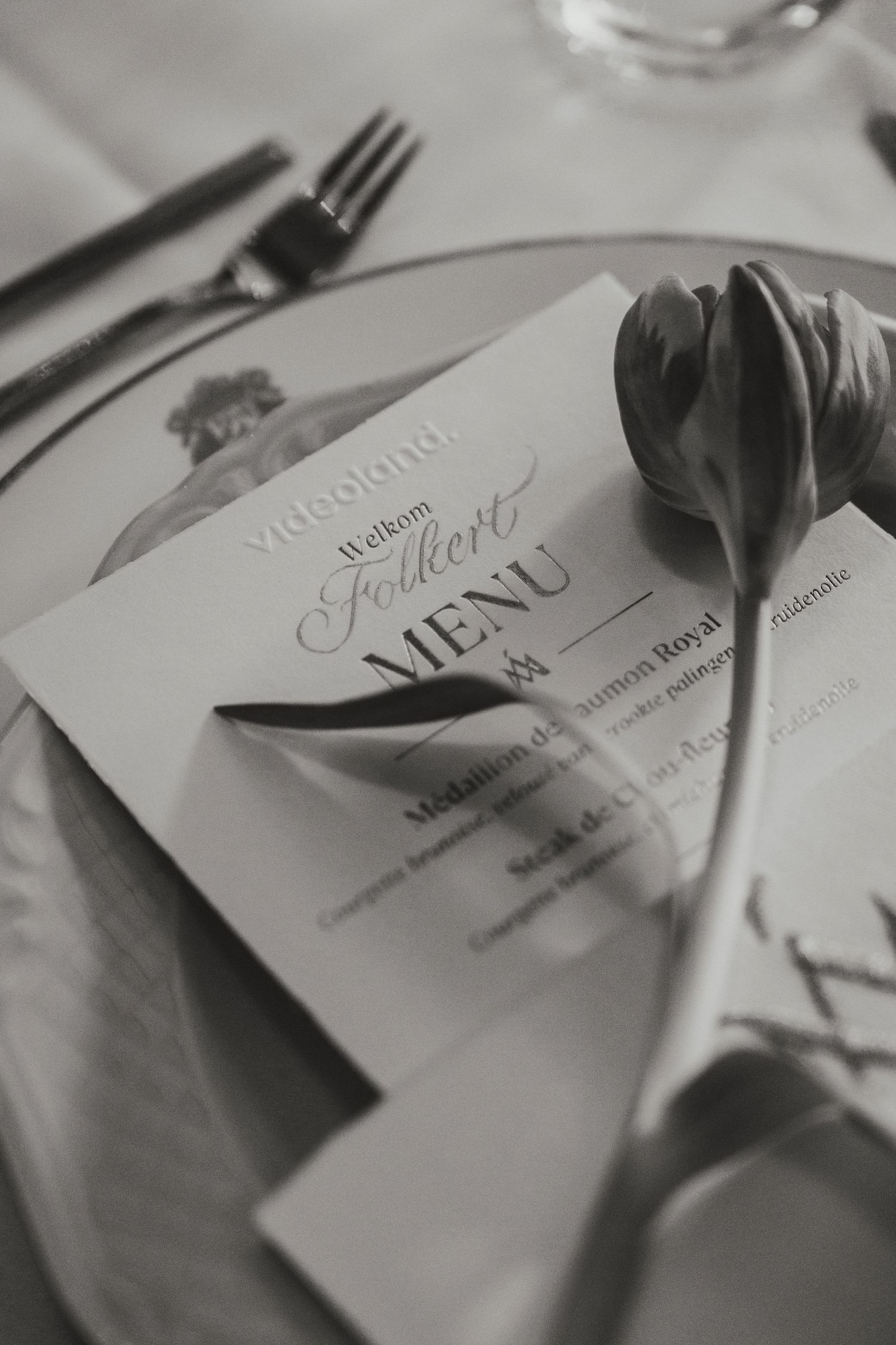 Black and white photo of an elegant place setting with a tulip, menu card, and cutlery at a formal dinner event.