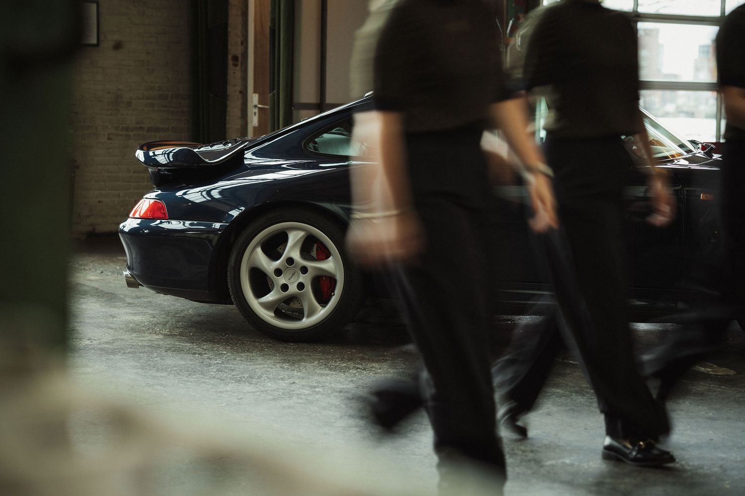 Black Porsche 911 GT3 parked in a garage with blurred figures walking past in the foreground.