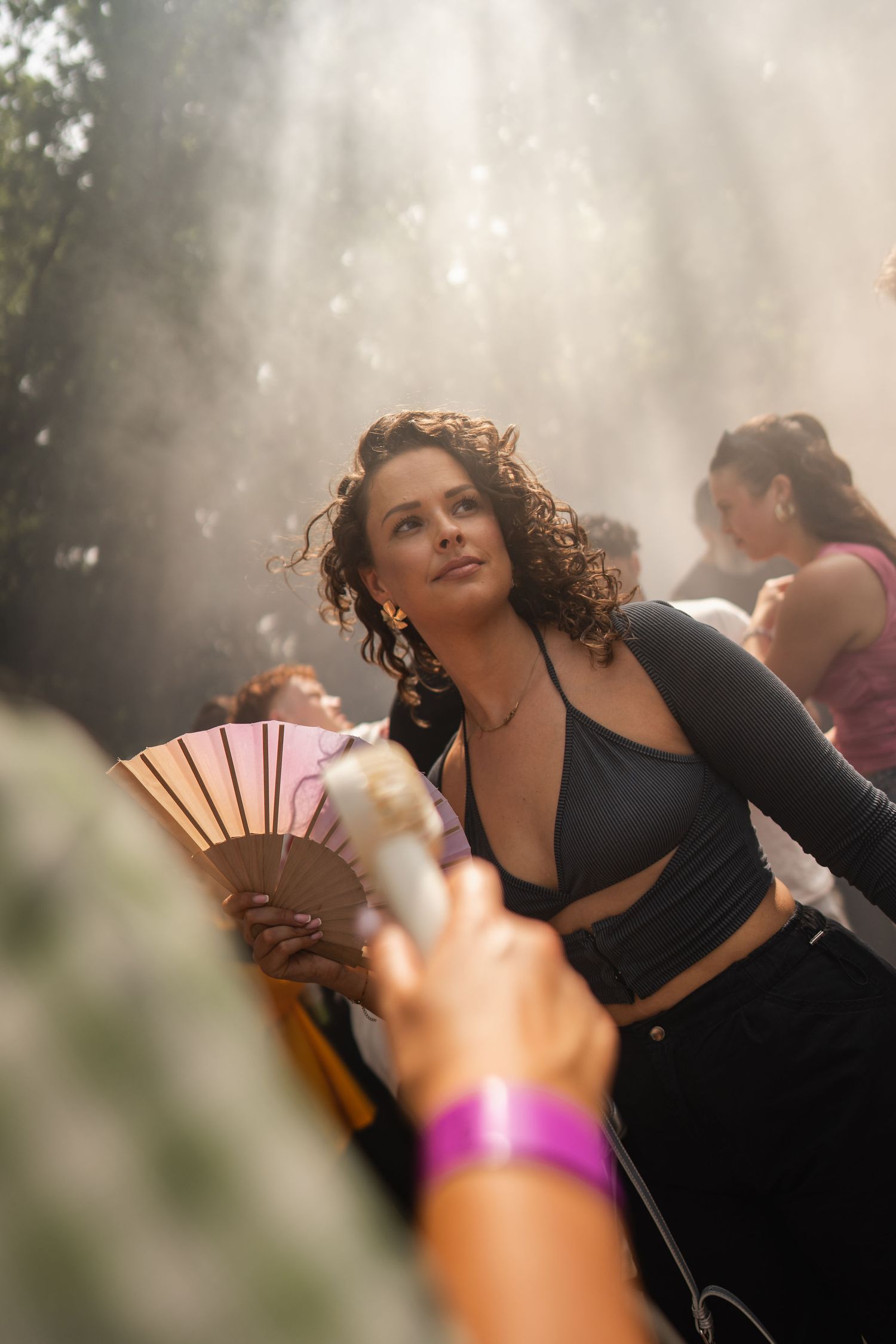 Young woman with curly hair holding a fan at an outdoor festival, surrounded by mist and sunlight rays.