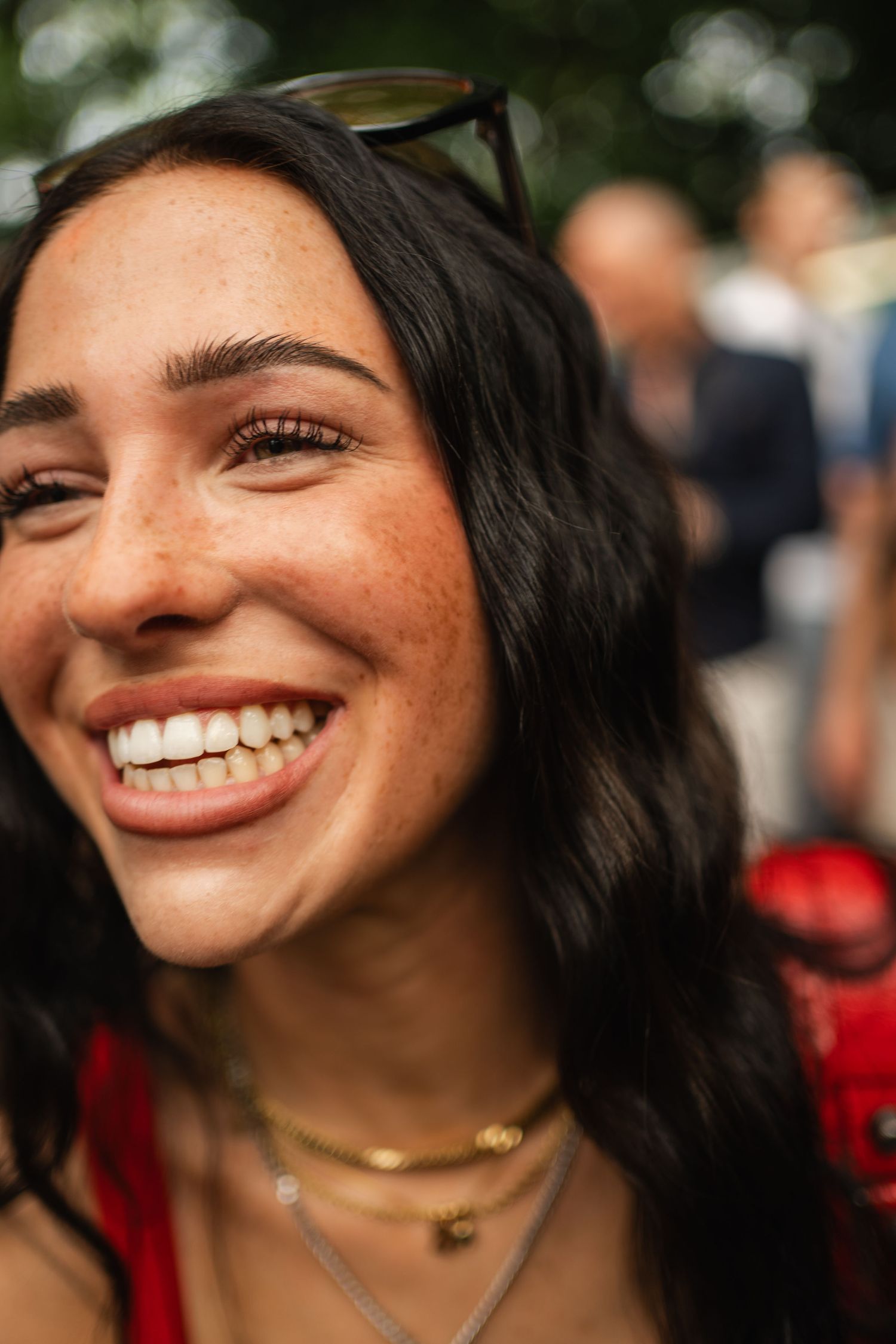 Smiling young woman with freckles, dark wavy hair, sunglasses on head, gold necklaces, wearing red at an outdoor event.