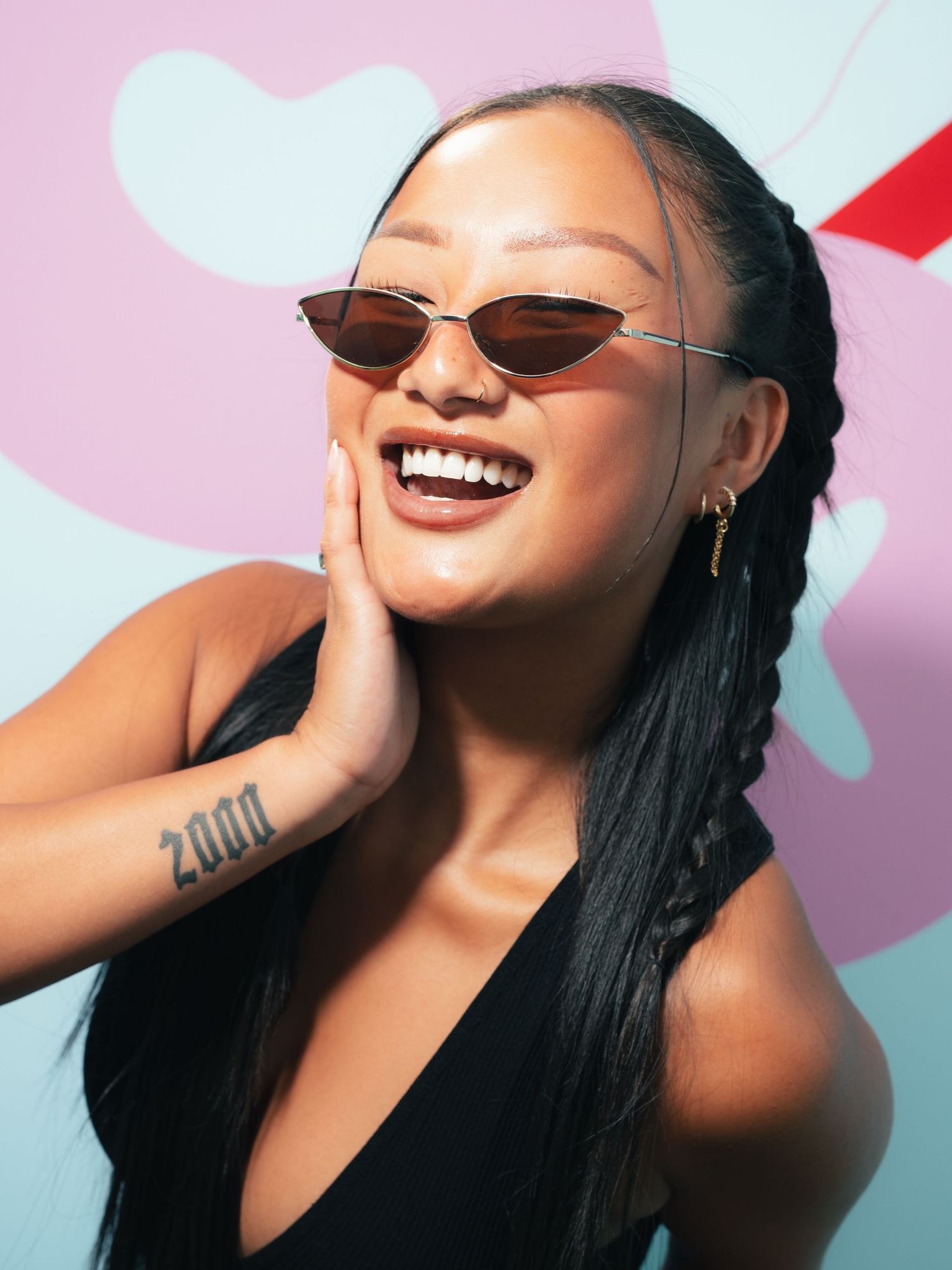 Smiling young woman with braids and small cat-eye sunglasses, hand on cheek, wearing a black top with a wrist tattoo.