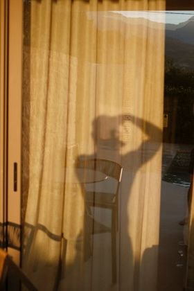 Shadow of a person silhouetted on sheer golden curtains by a window with mountain views at sunset.