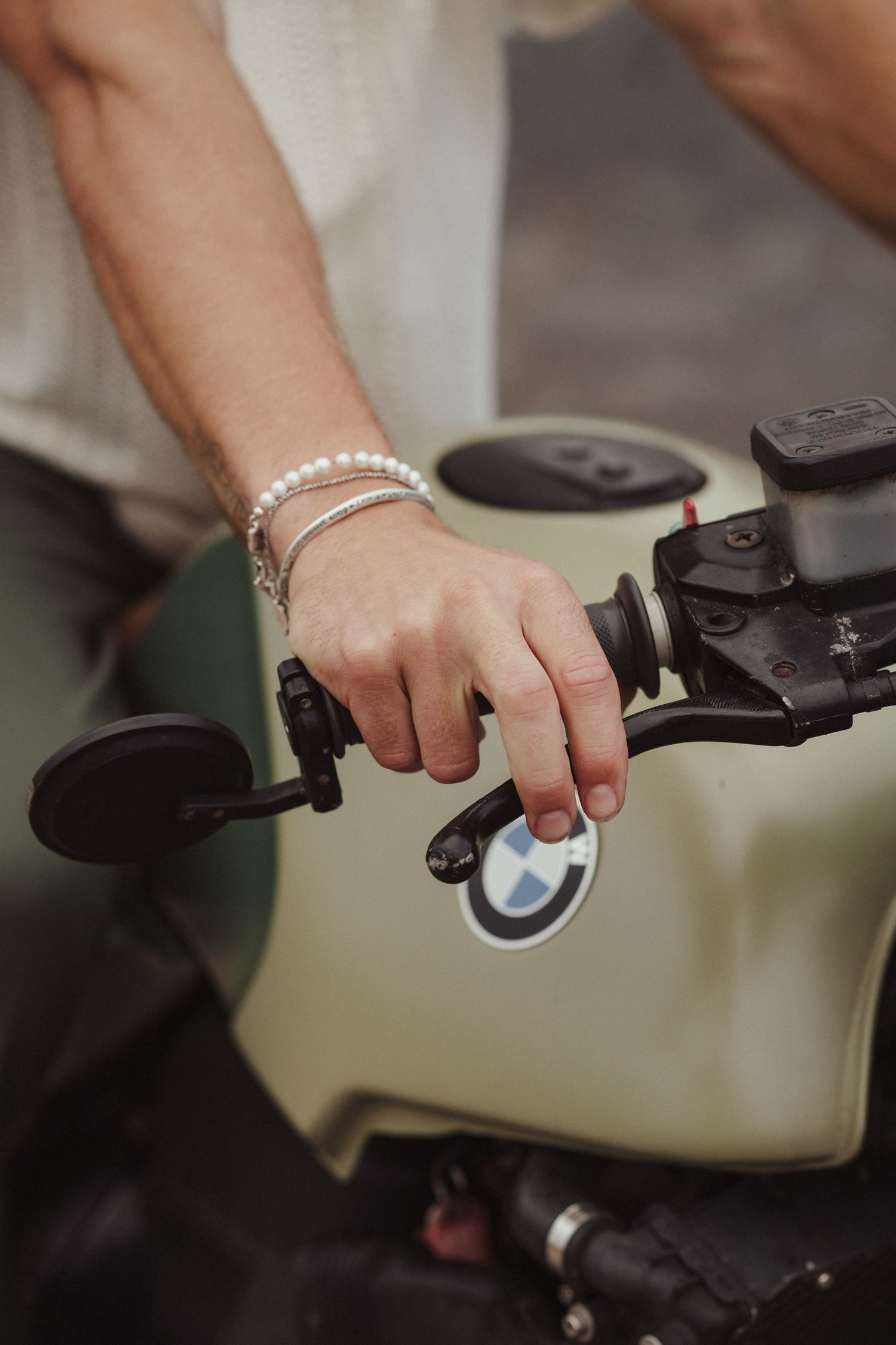 Close-up of a hand with pearl bracelet gripping a BMW motorcycle handlebar, olive green fuel tank visible.
