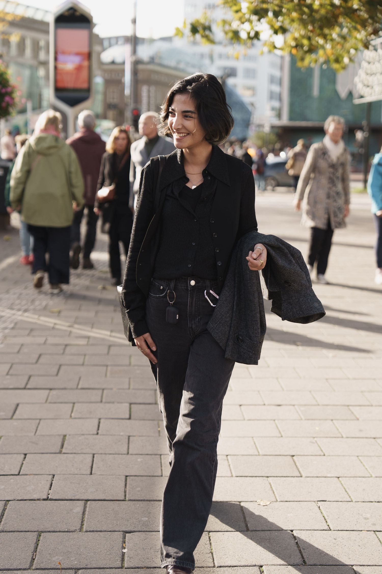 A smiling woman in black outfit walks confidently on a sunny urban sidewalk with people in the background.