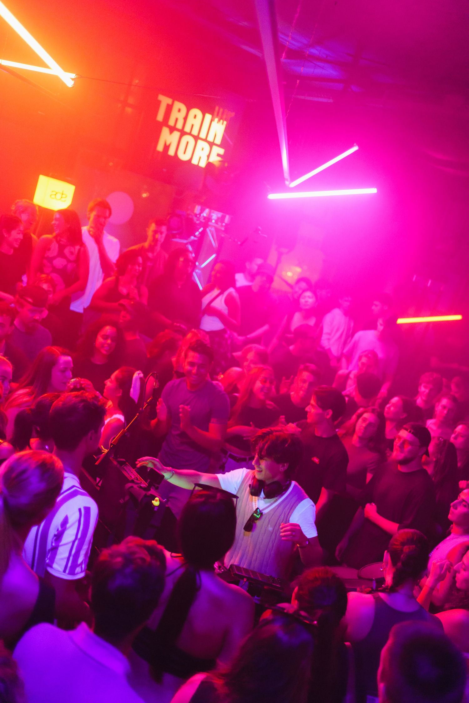 Crowd of partygoers dancing under pink and orange neon lights at a nightclub with a Train More sign.