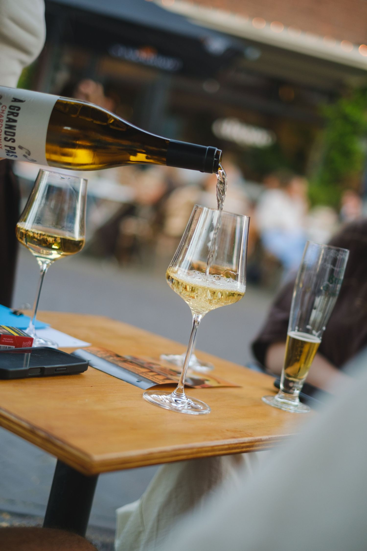 White wine being poured into elegant stemmed glasses on a wooden table at an outdoor restaurant terrace.