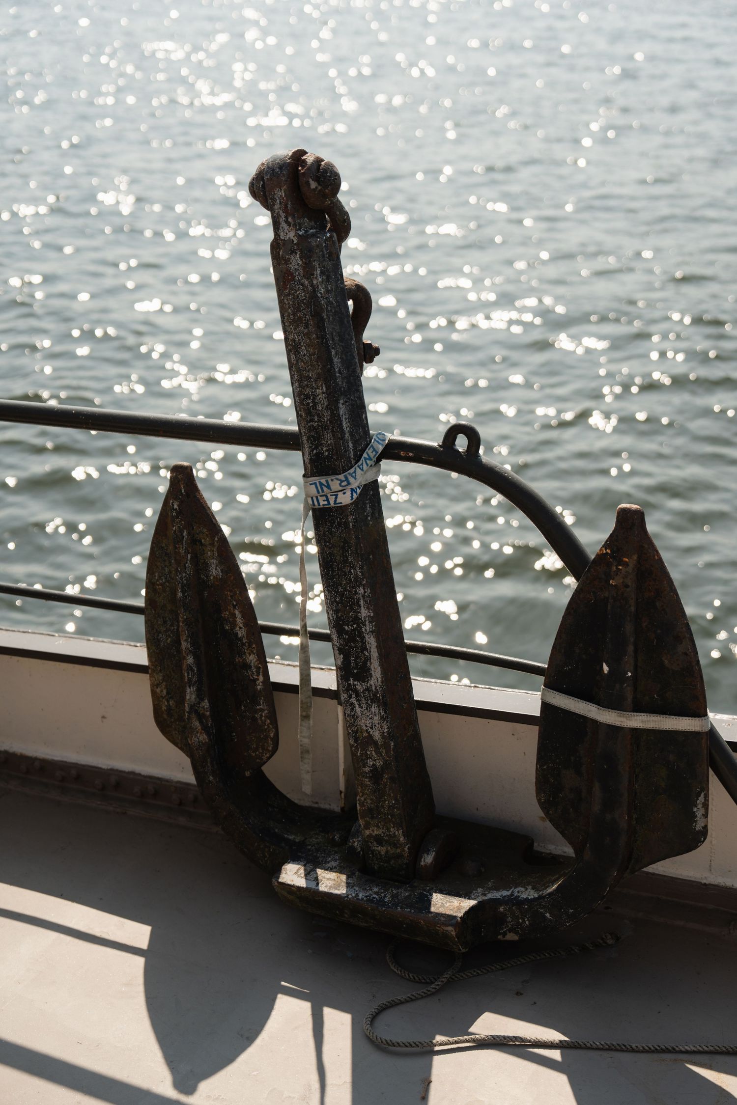 A weathered black iron anchor rests on a boat railing with sunlight sparkling on the water in the background.