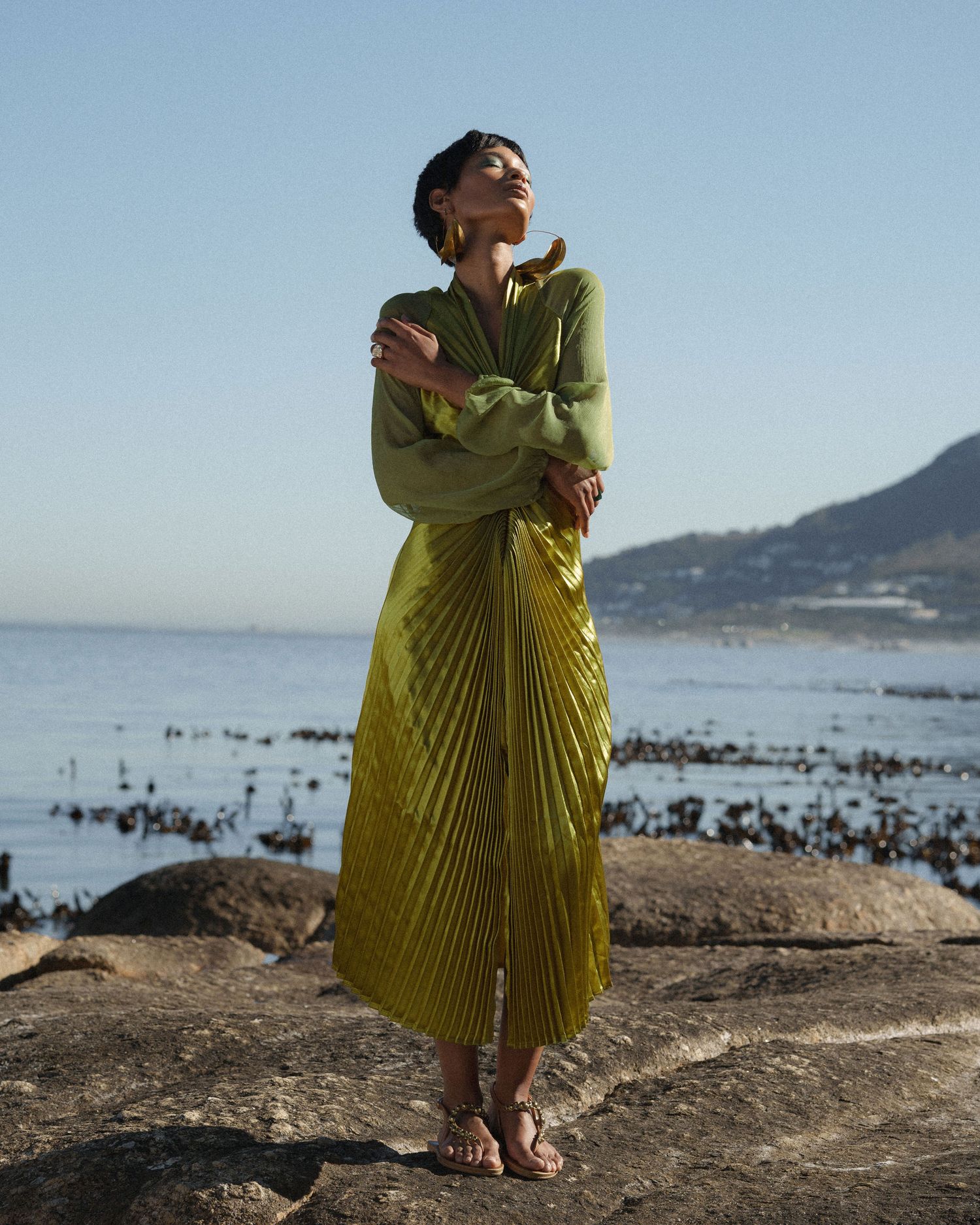 Woman in chartreuse pleated midi dress standing on rocks by the ocean with mountains in background.