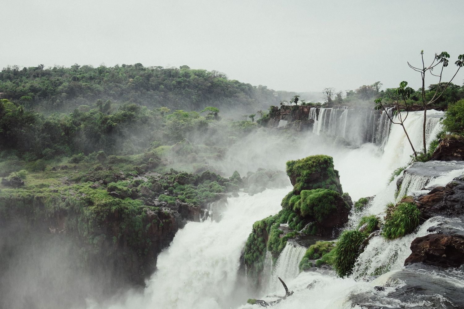Majestic waterfalls cascade over moss-covered rocks into misty rapids, surrounded by lush tropical forest.