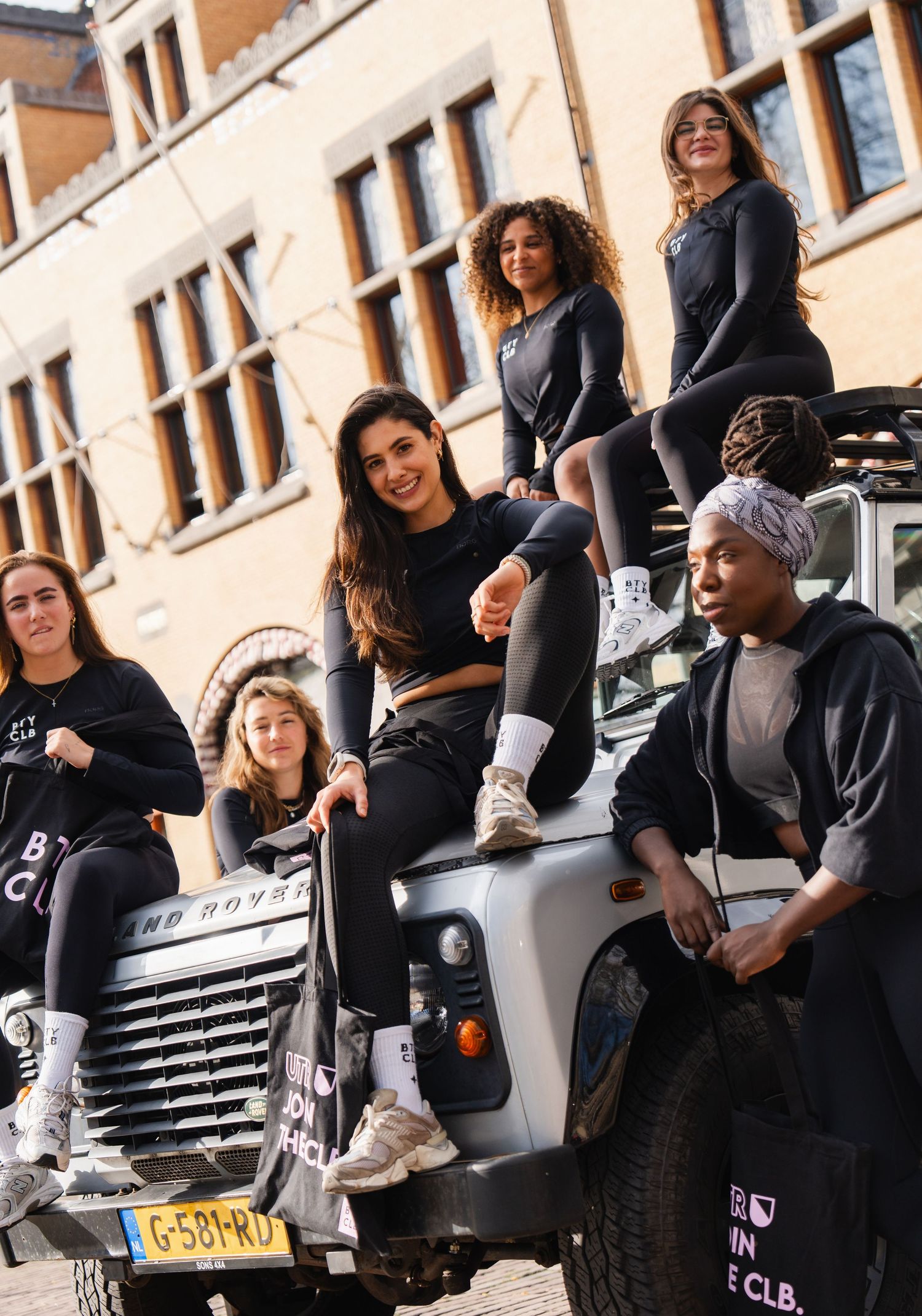 Seven diverse women in black athletic wear posing on a Land Rover SUV on a city street.