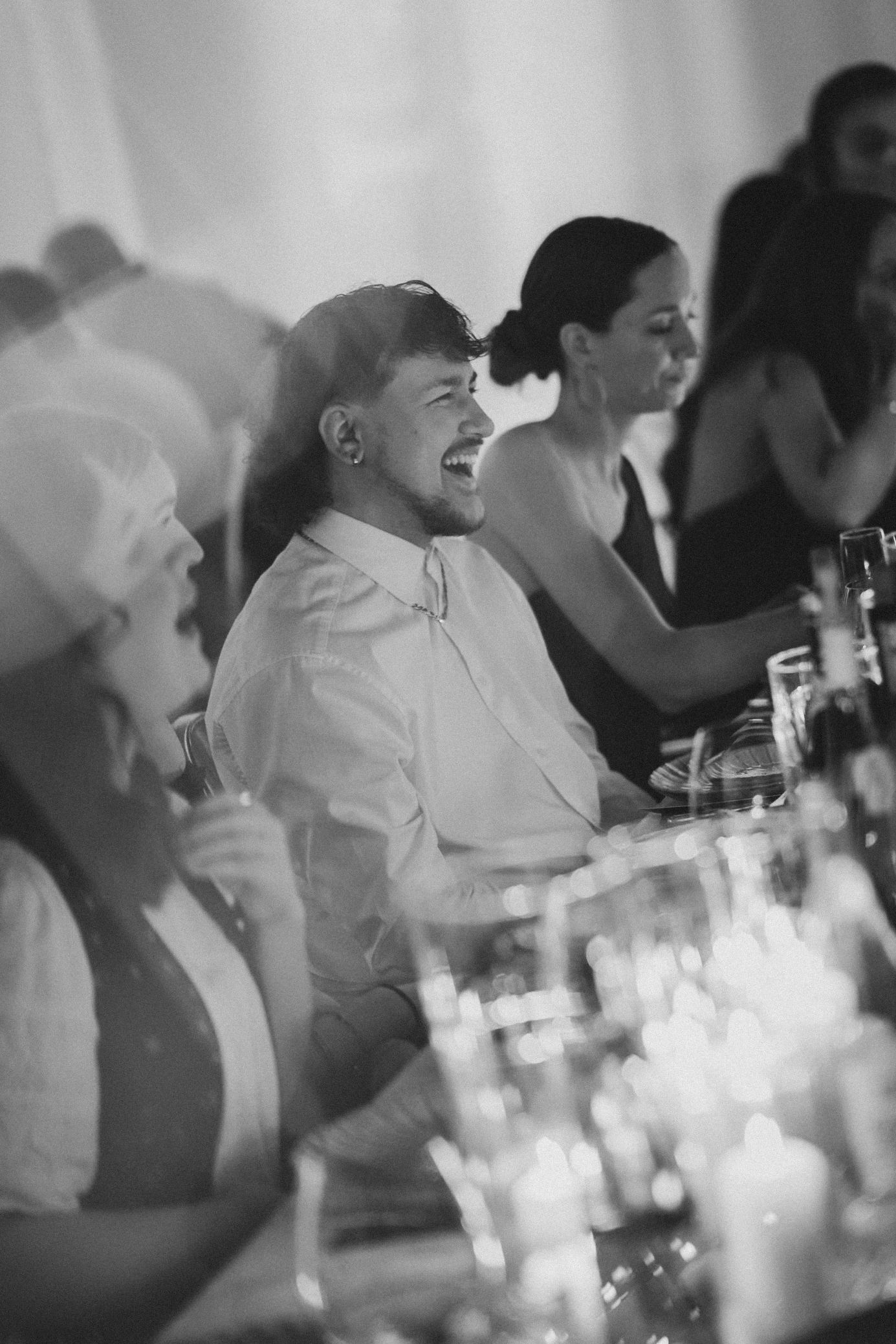 Black and white photo of laughing wedding guests seated at a candlelit reception dinner table.