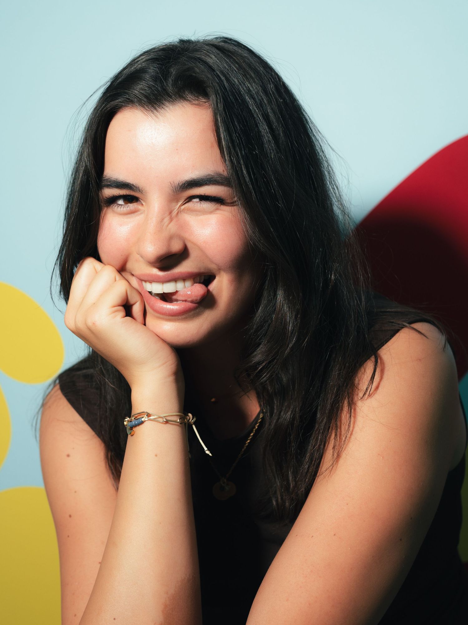 Young brunette woman in a black top laughing with hand near face, wearing a gold bracelet indoors.