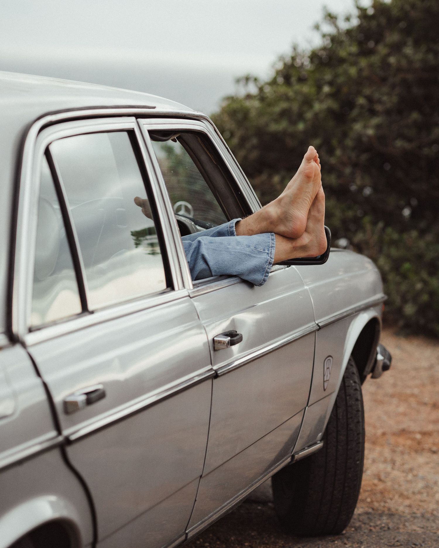 Bare feet and jeans legs hanging out of a silver vintage car window with green shrubs behind.