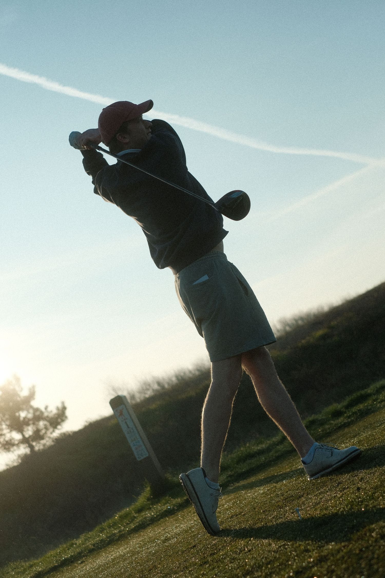 Golfer completing a follow-through swing on a golf course at sunset, wearing a red cap and khaki shorts.
