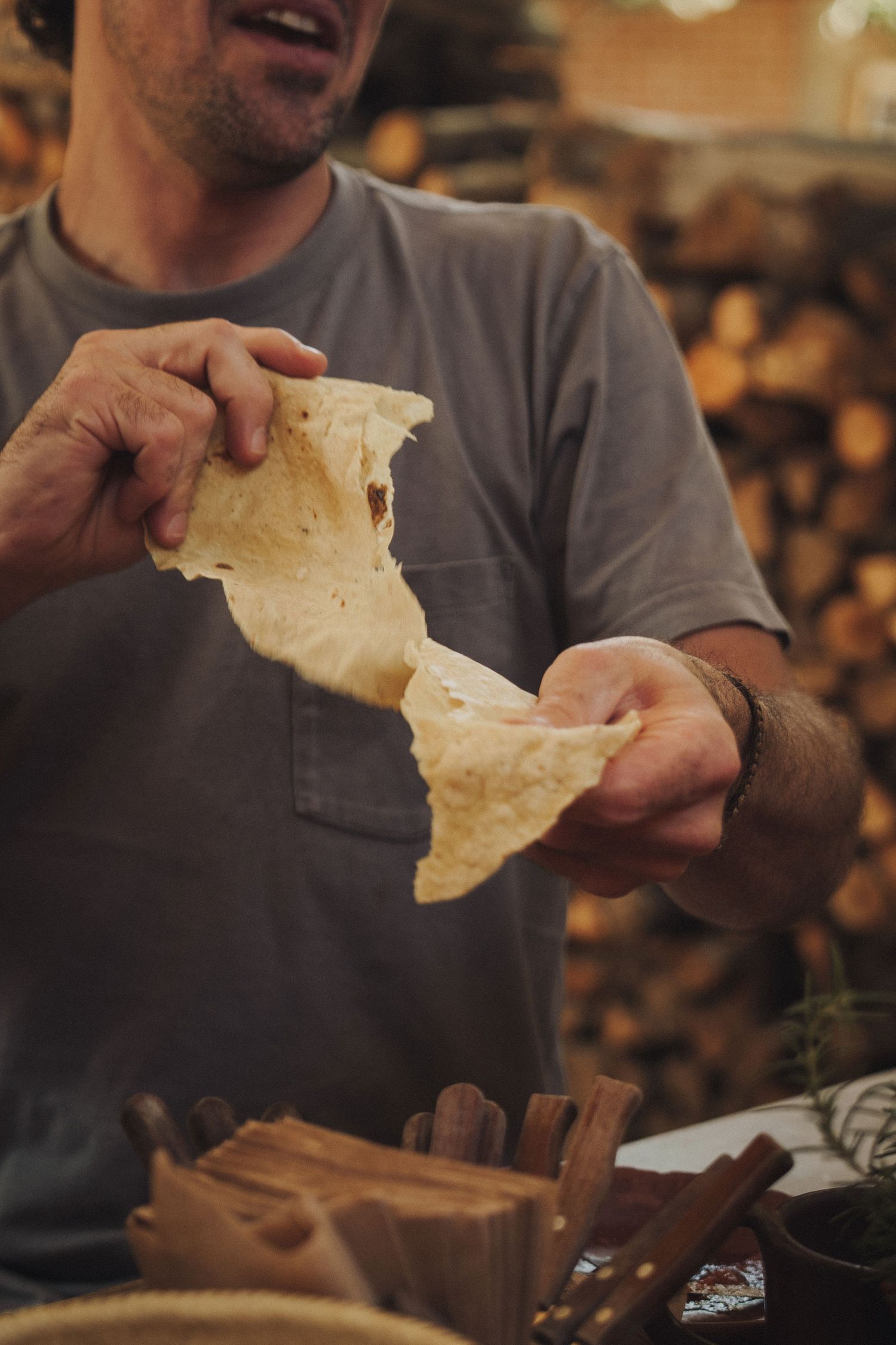 Man tearing fresh flatbread by hand near a rustic wood pile, showcasing artisan bread making.