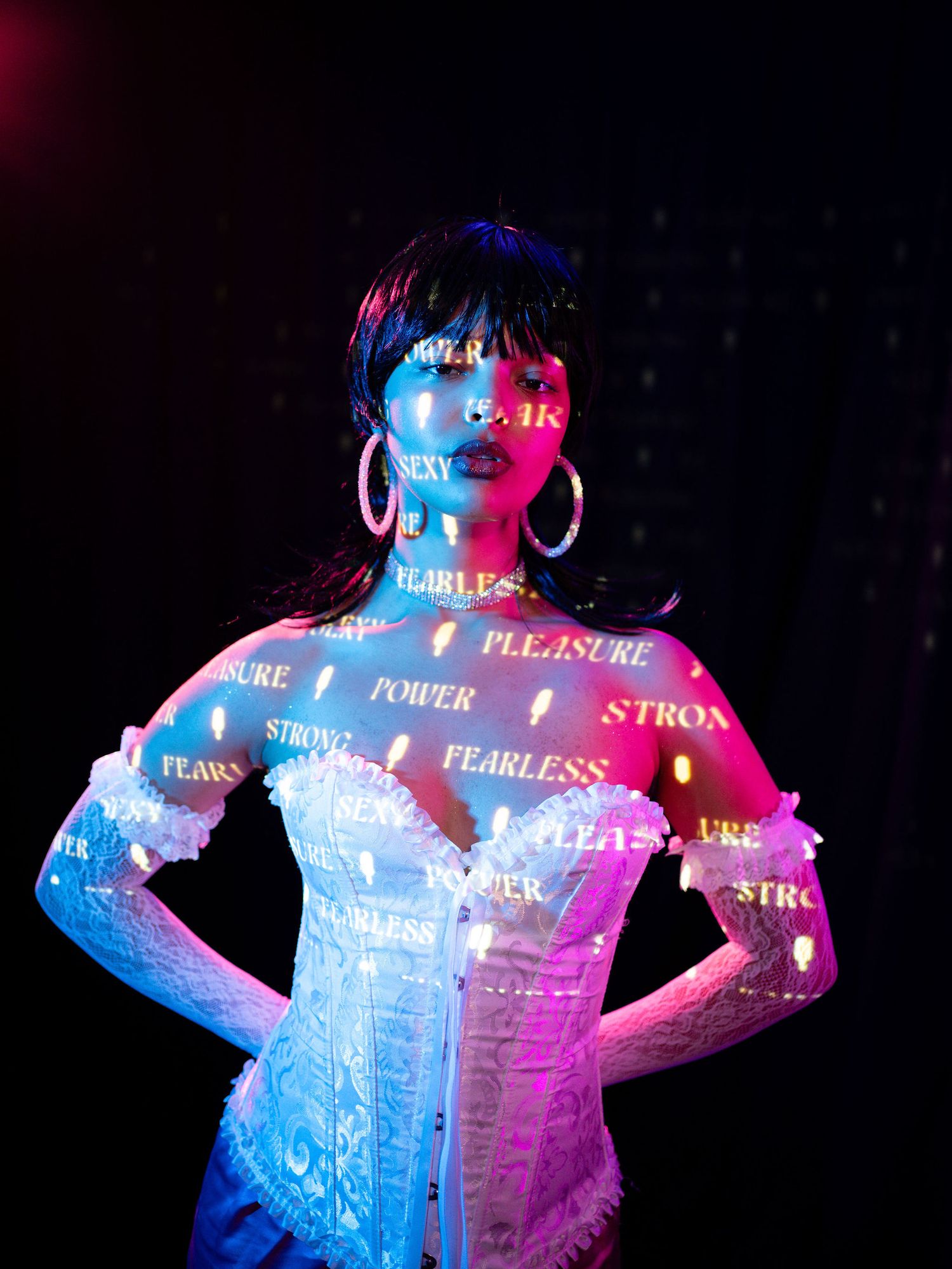 Woman in white corset with empowering words projected in neon pink and blue light against dark background.