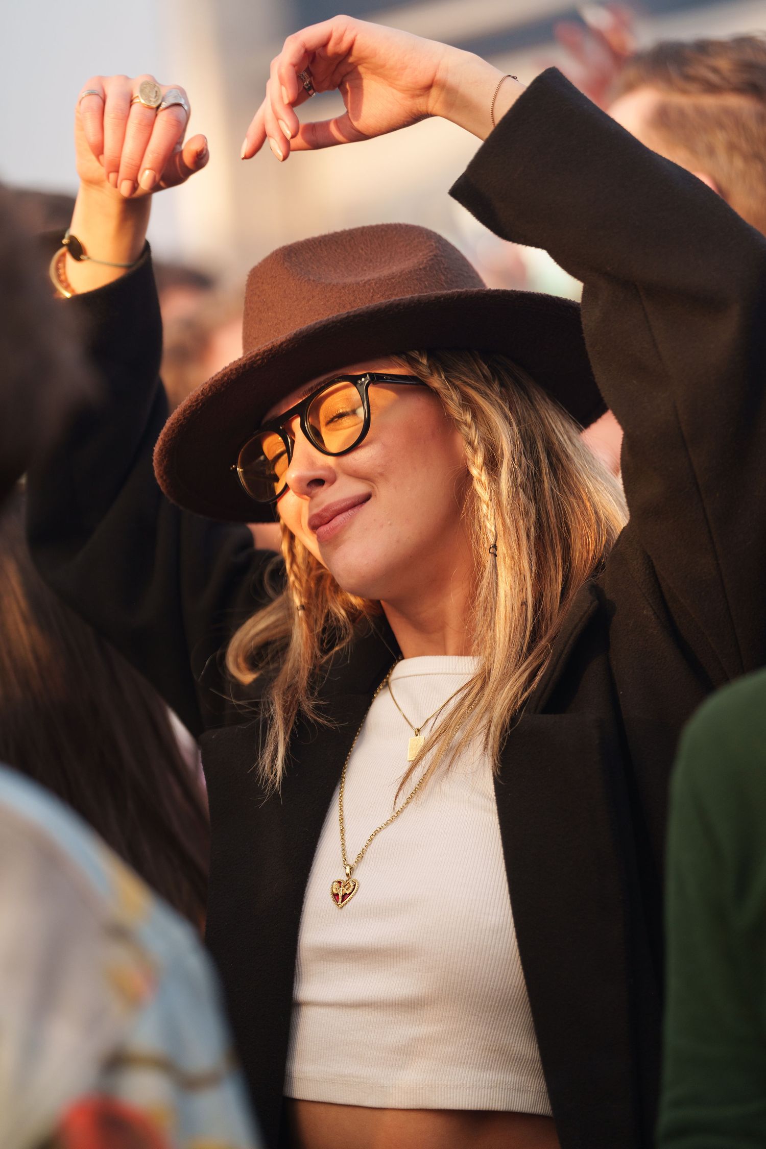 Woman dancing at festival wearing brown wide-brim hat, sunglasses, white crop top, and black blazer with gold necklace.